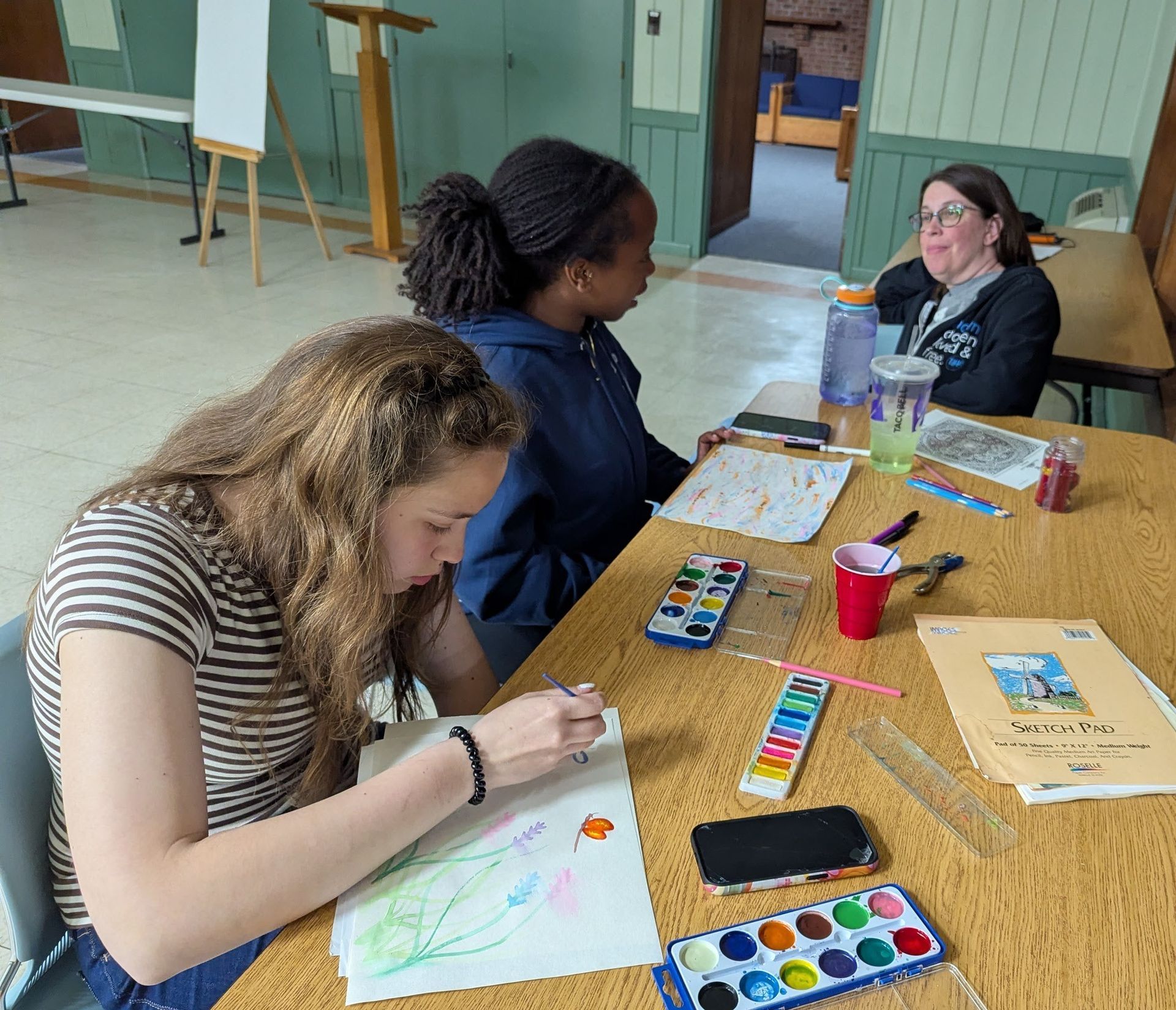 Three people coloring at a table in a bright room with art supplies and drinks.