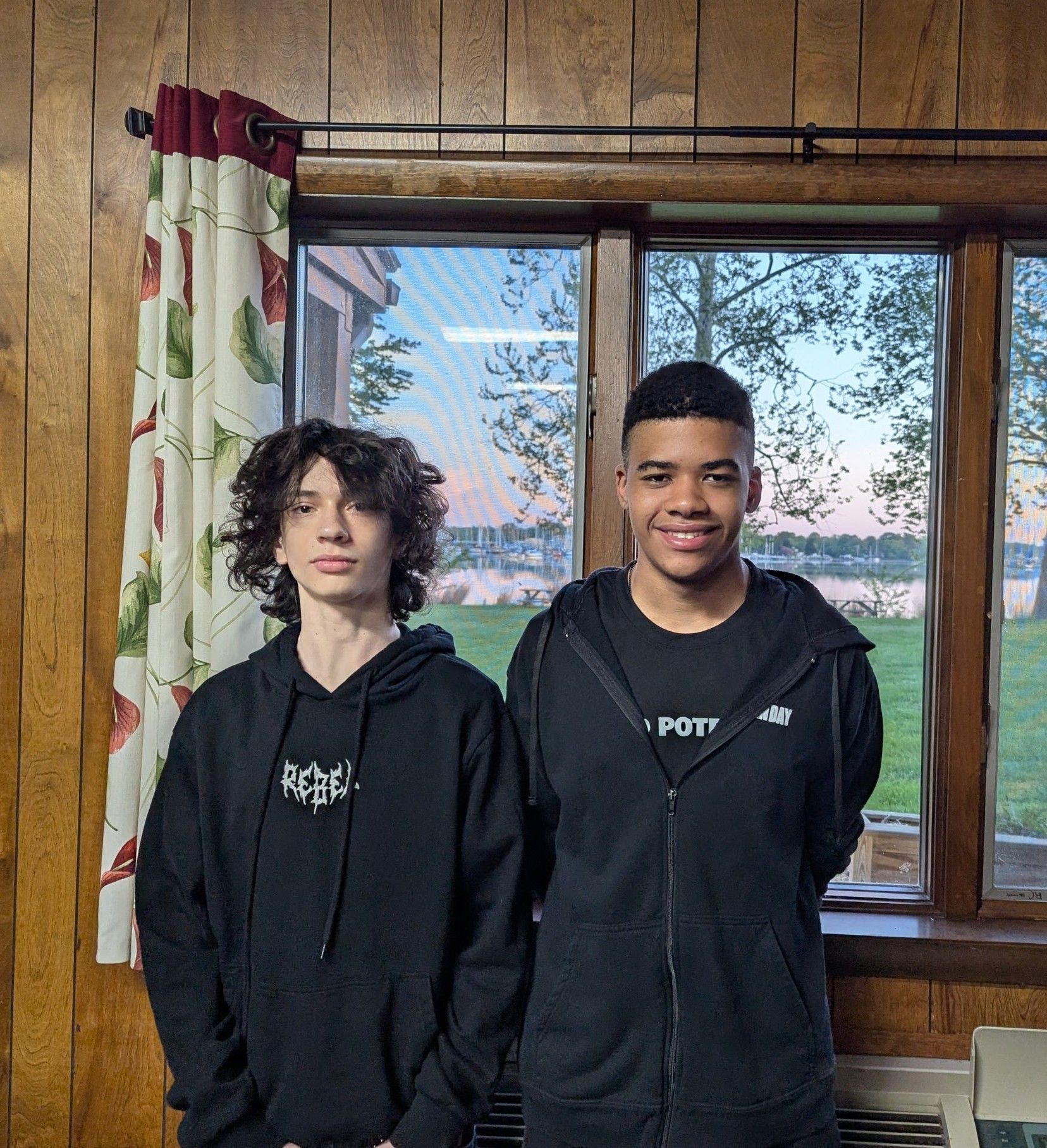 Two friends in black hoodies standing indoors by a window with a scenic view.