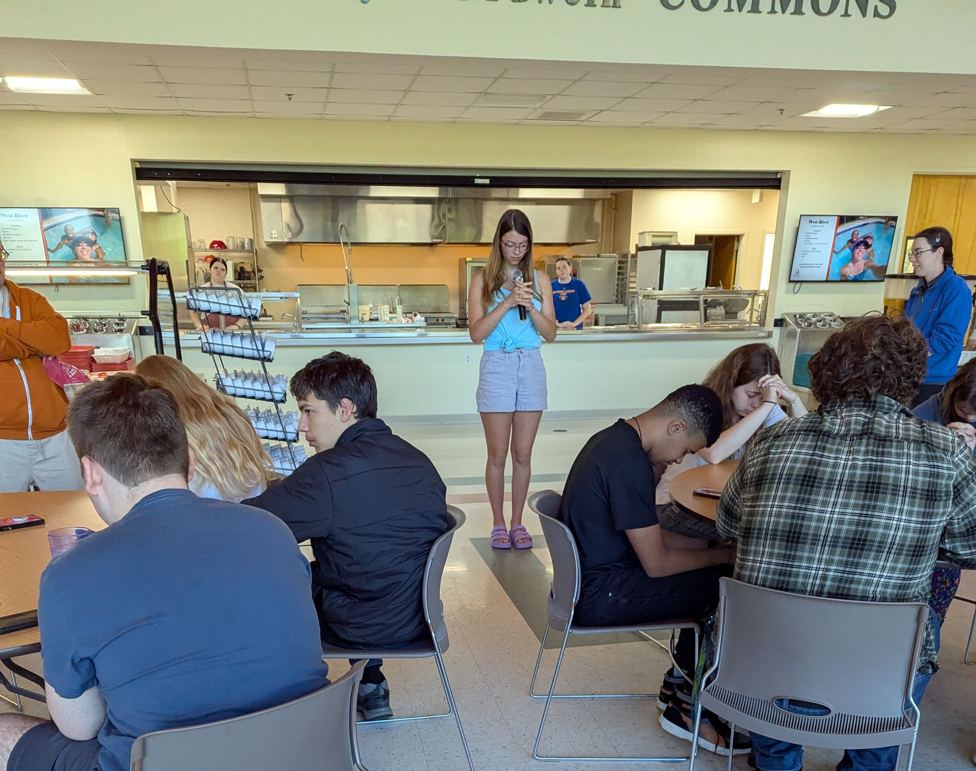One youth saying grace in a cafeteria while several youth bow their heads.
