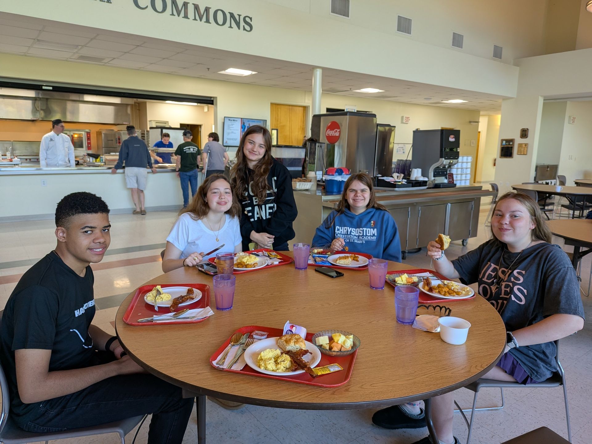 Youth seated around a cafeteria table with trays of food, smiling in a commons area