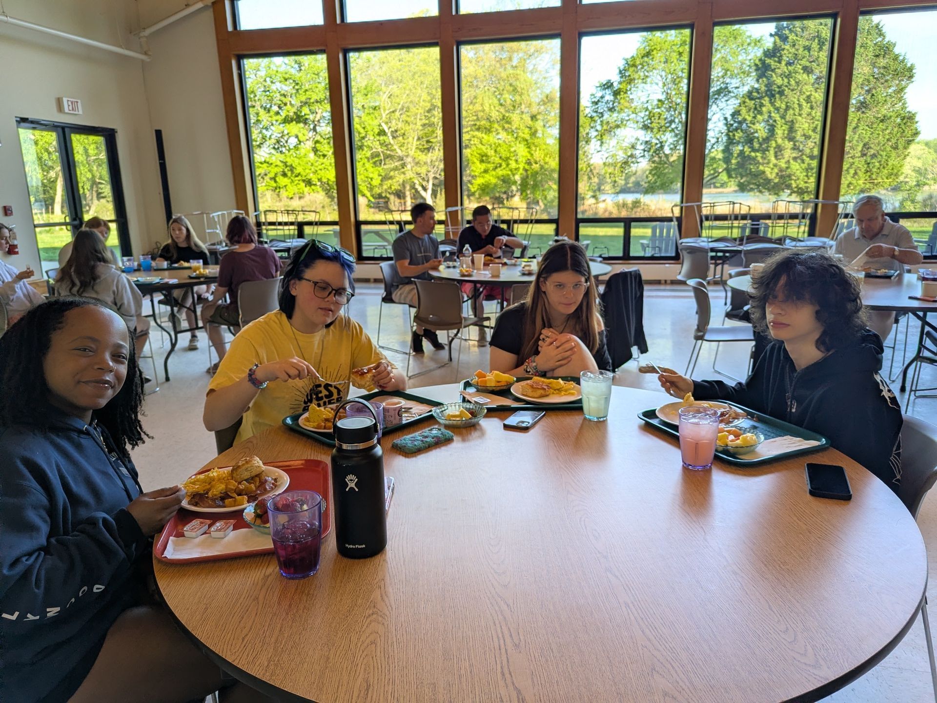 People seated around a table in a bright cafeteria, eating lunch and chatting near large windows.