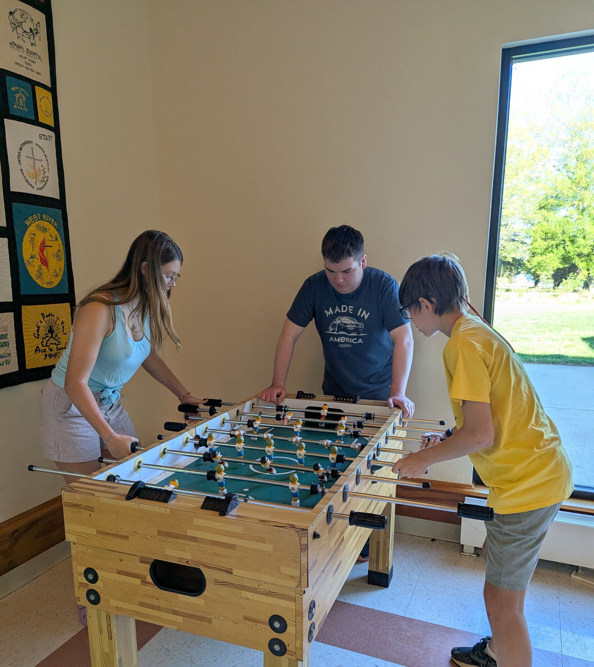 Three people playing foosball indoors near a bright doorway