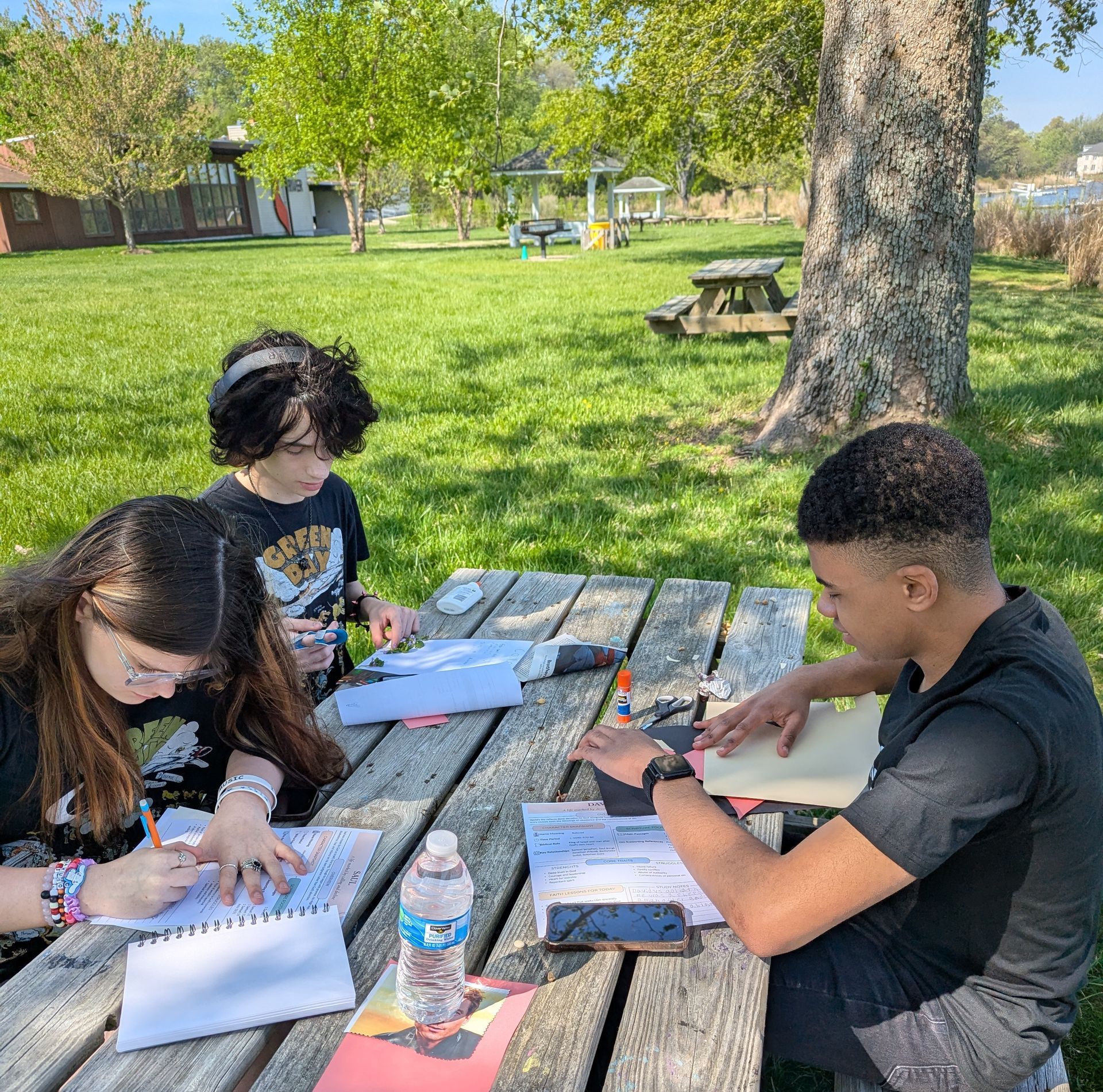 Three students writing at a picnic table in a sunny park, with papers, notebooks, and a water bottle.