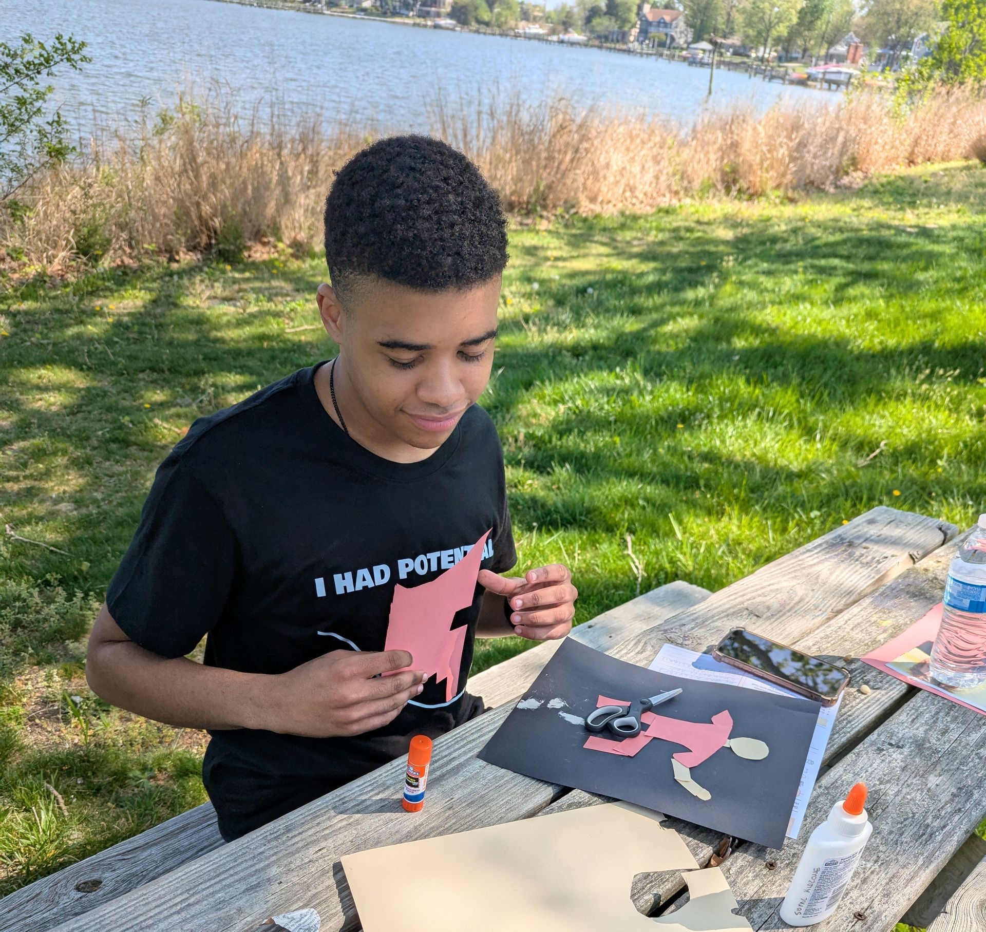 A youth painting a pink cutout at a picnic table beside a lake on a sunny day
