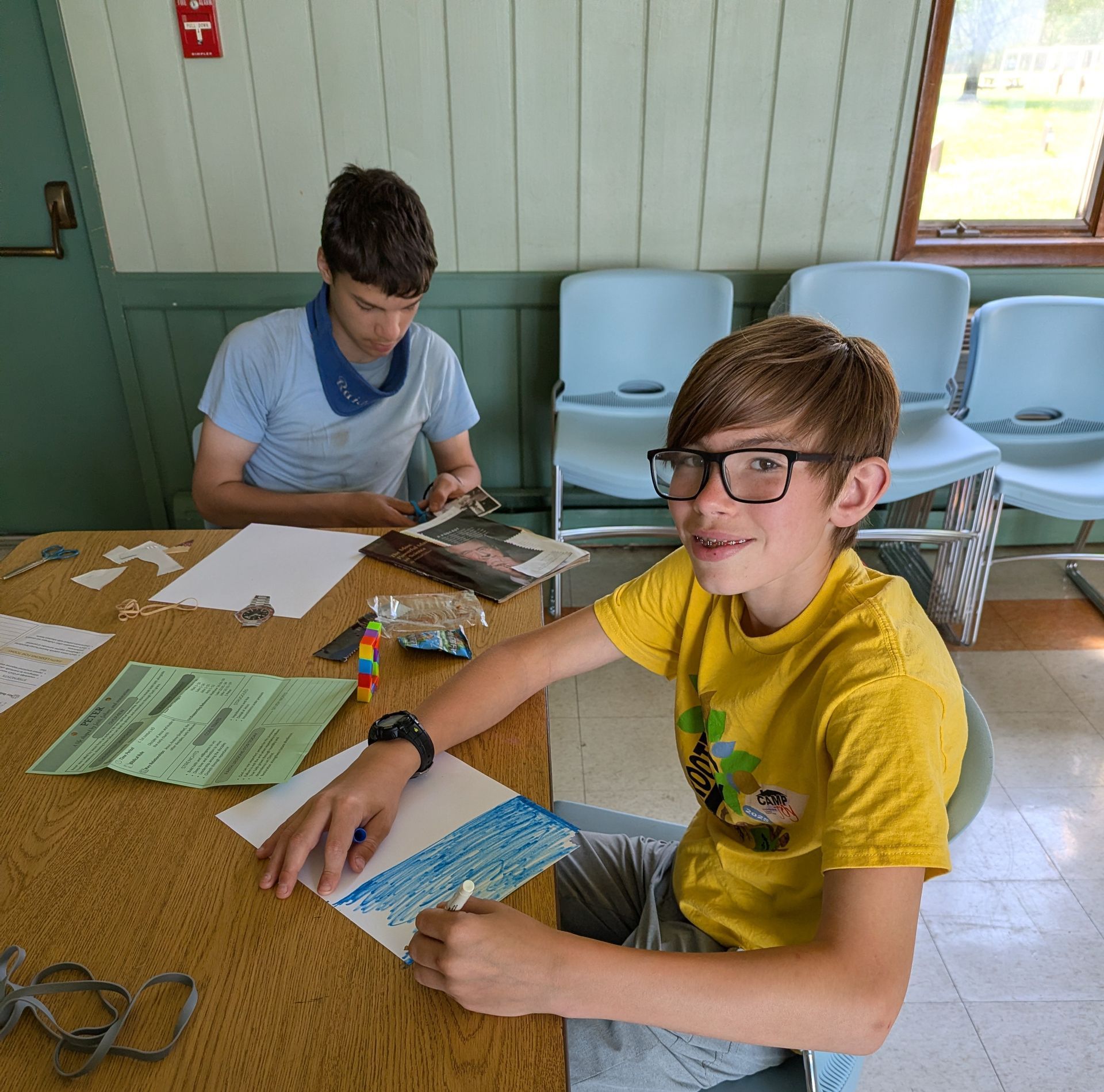 Two students at a table with papers, markers, and a laptop in a classroom-like room