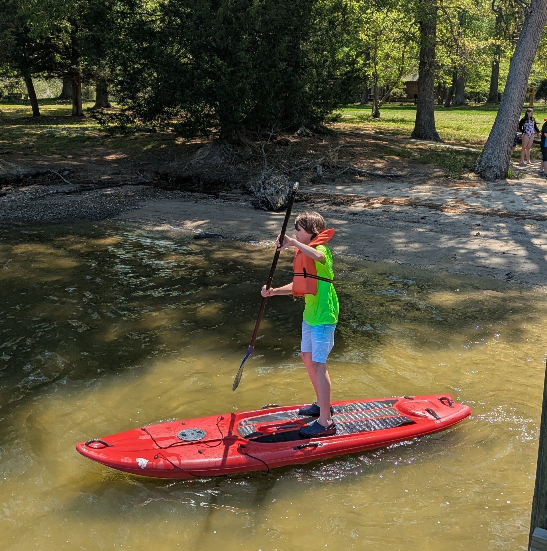 A youth paddleboarding on a red board in shallow water near a wooded shore