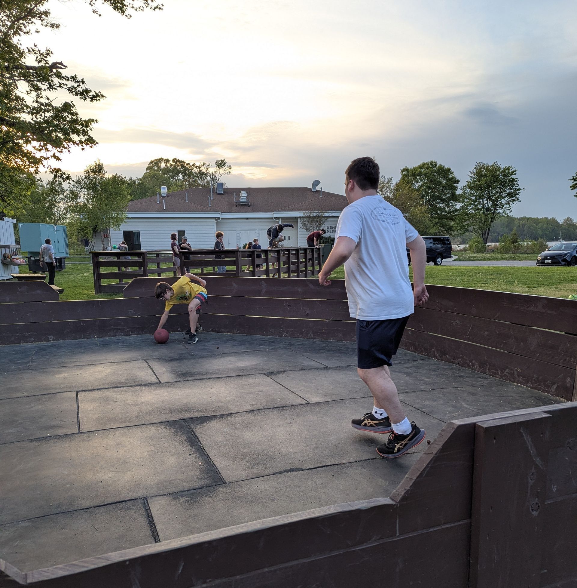 Two people playing on a small outdoor skatepark at sunset, with trees and a pavilion in the background.