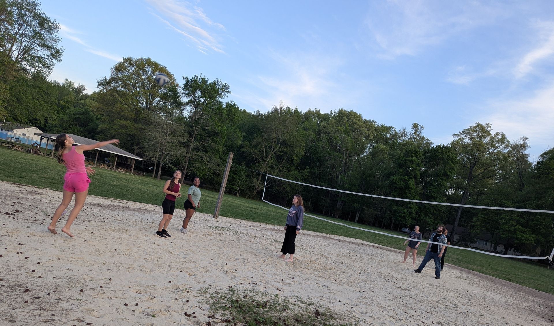 People playing volleyball on a sandy court outdoors with trees and a blue sky in the background