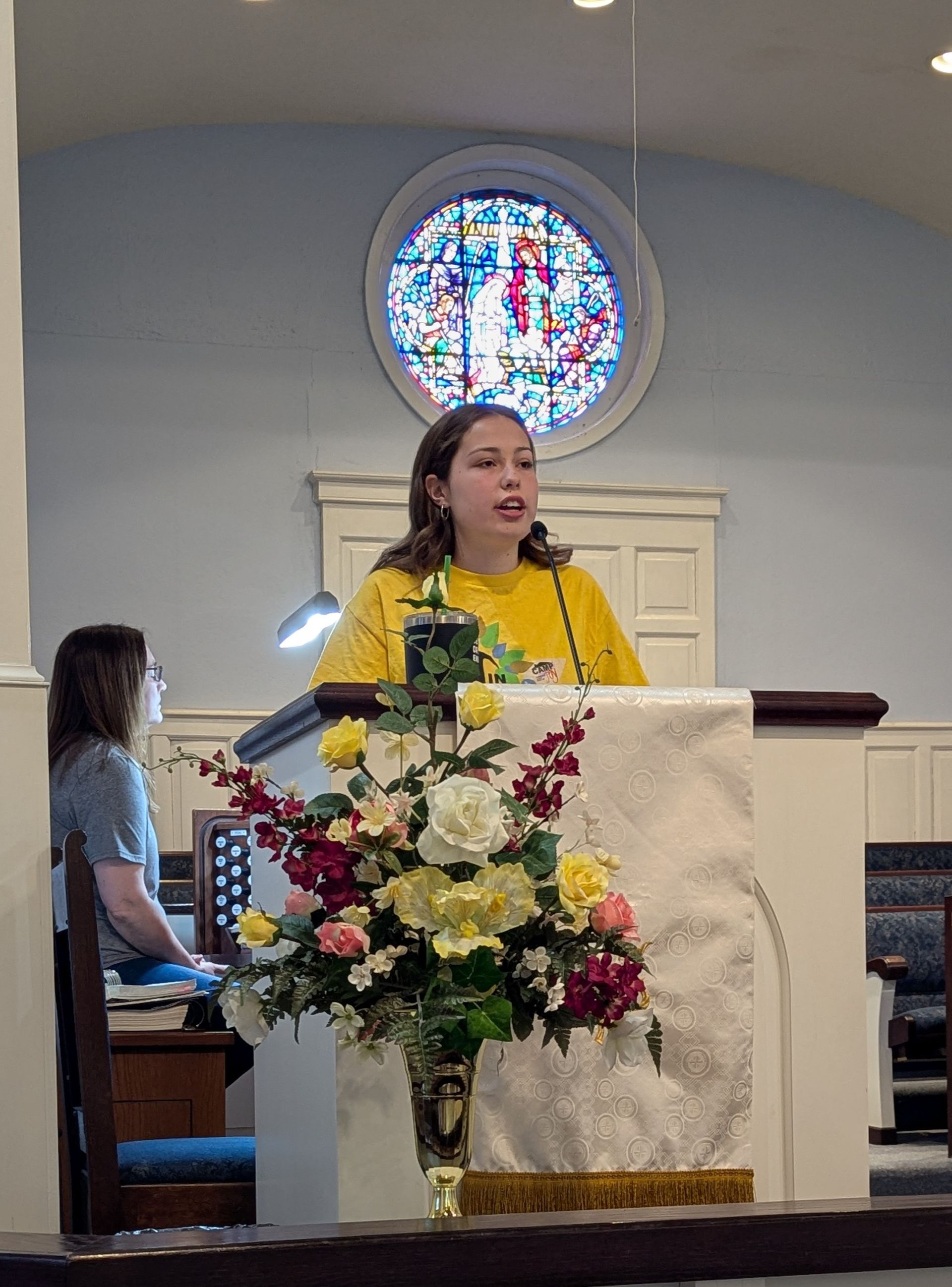 Youth speaking at a church pulpit with a floral arrangement and stained-glass window behind her.