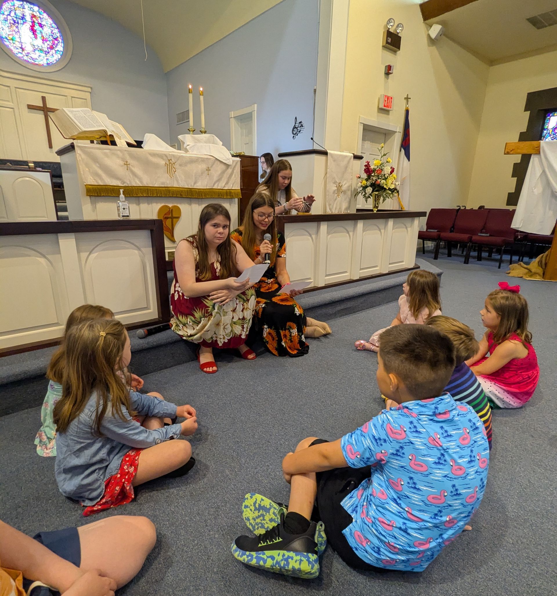 Children sitting on the floor listen to a youth in a church sanctuary near the altar.
