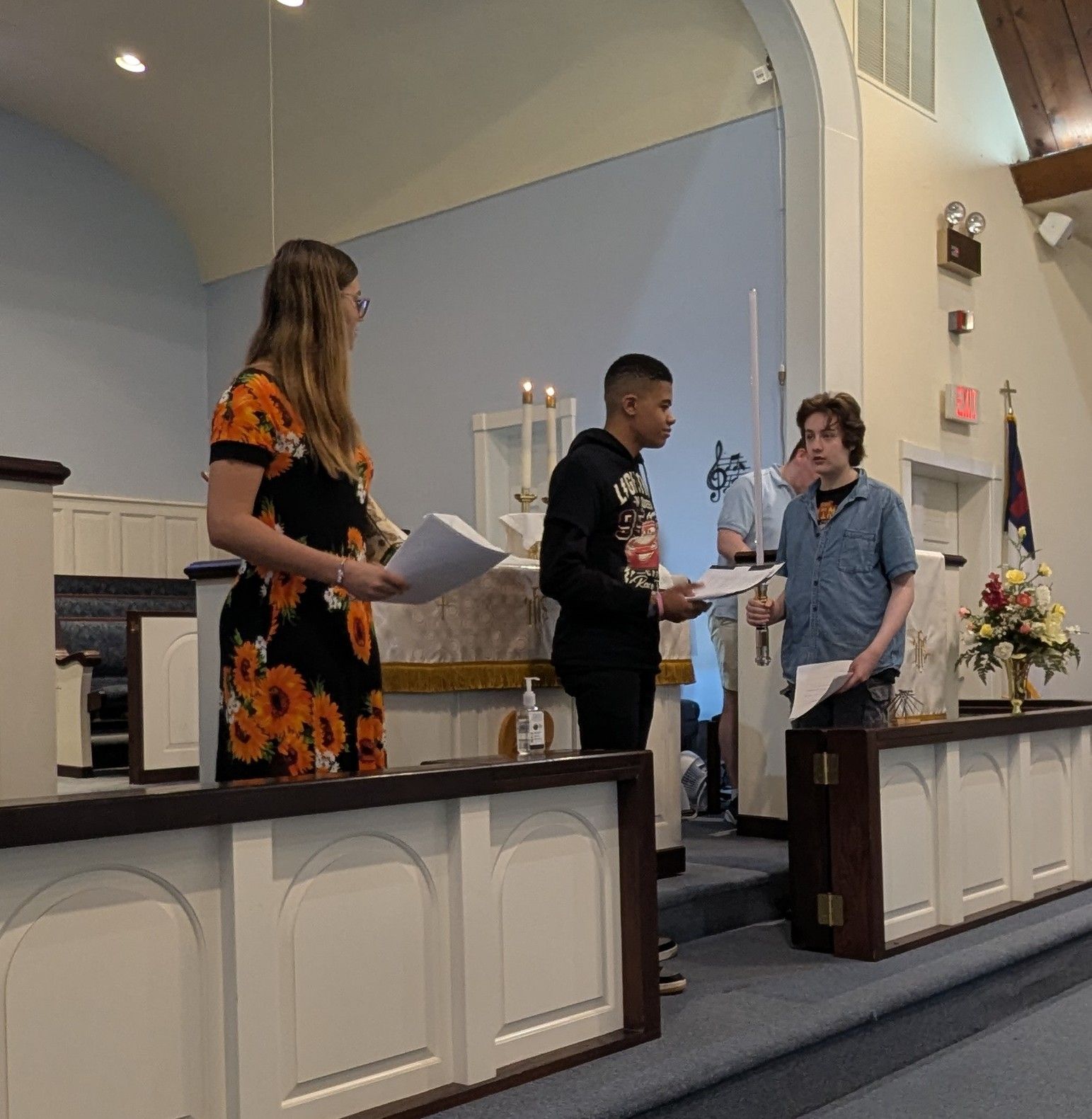 Three people stand at a church altar during a ceremony, one receiving a document.