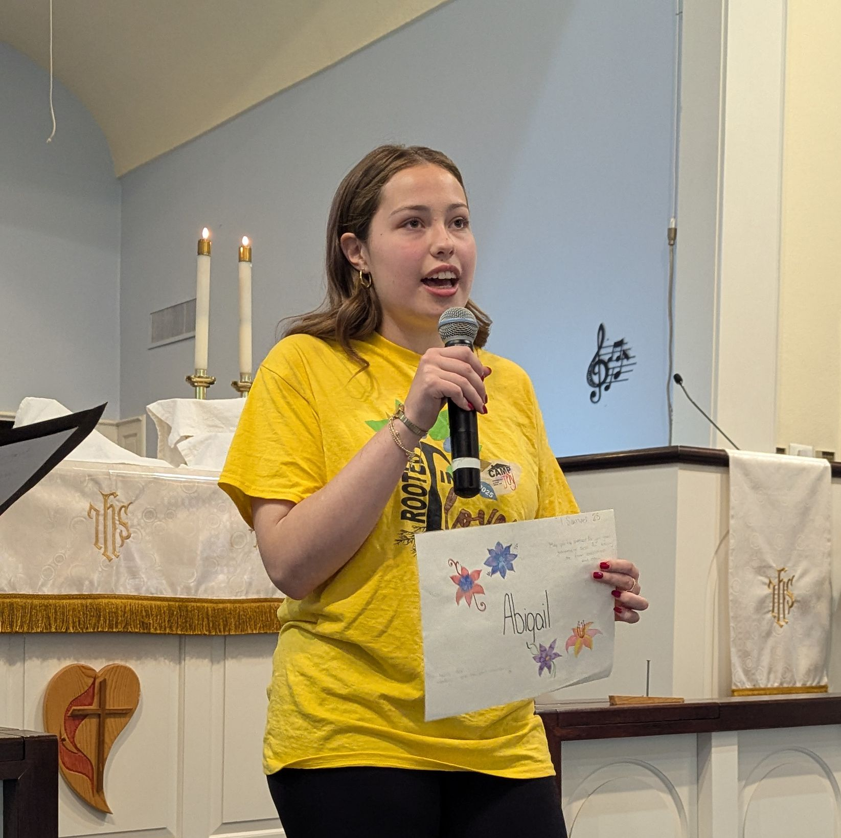 Youth in yellow shirt speaking into a microphone at a church altar while holding a decorated paper.