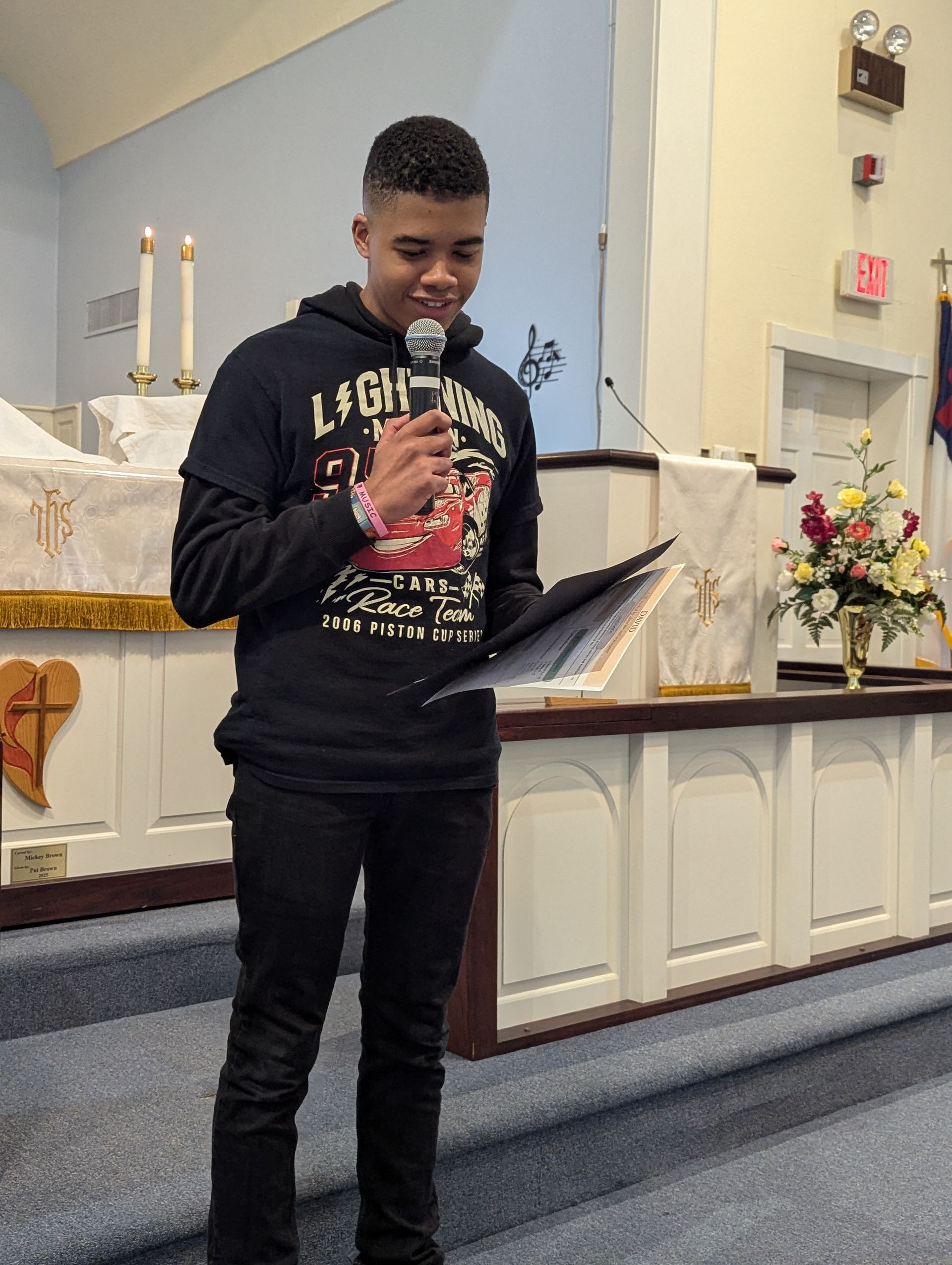 Youth reading from a folder at a church altar, wearing a black sweatshirt and standing near floral arrangements.