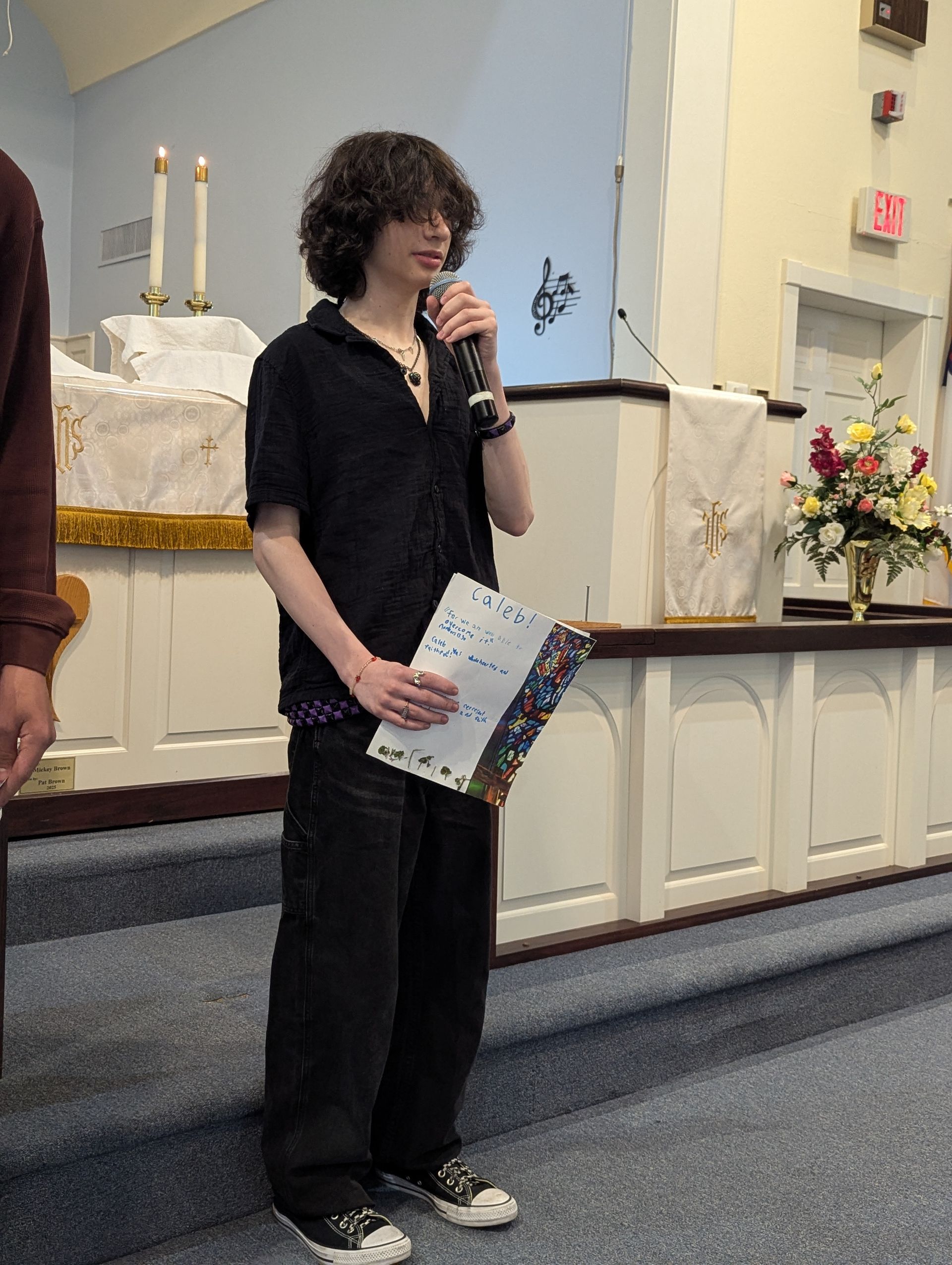 Youth speaking into a microphone at a church altar, holding papers, with candles and flowers nearby.