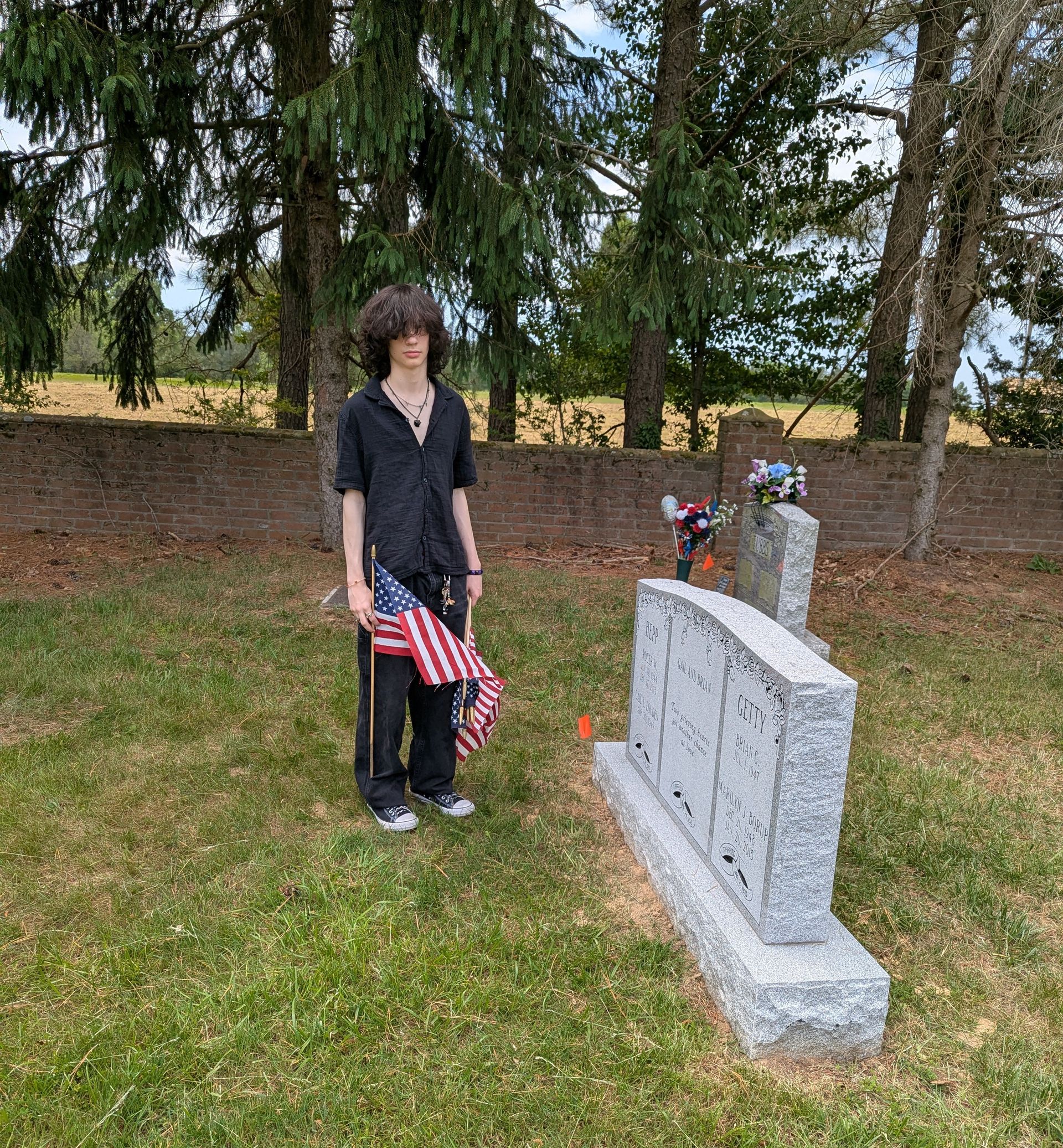 A teen stands beside a gravestone holding an American flag in a grassy cemetery.