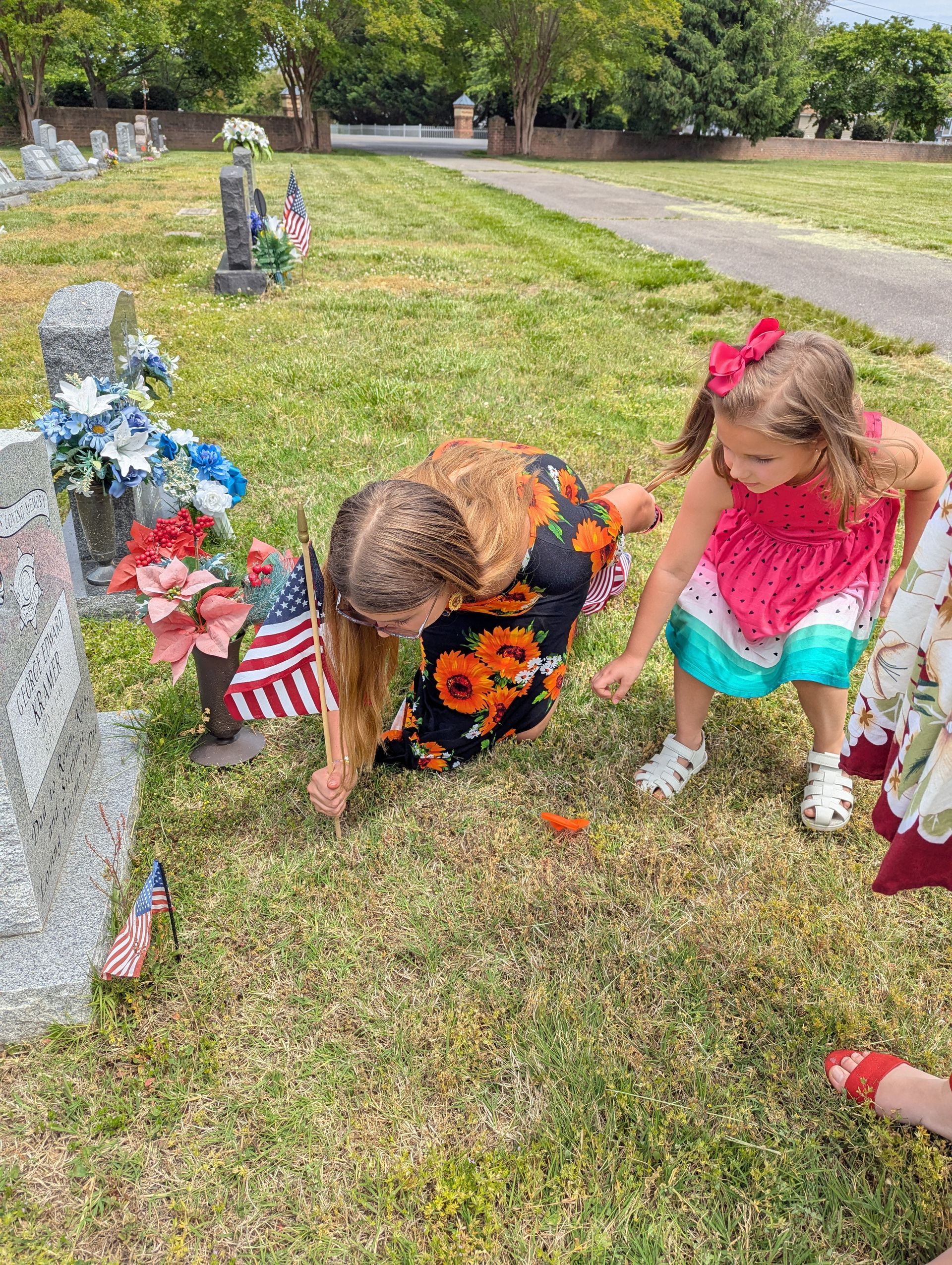 Children kneel at a gravesite decorated with flowers and small American flags.