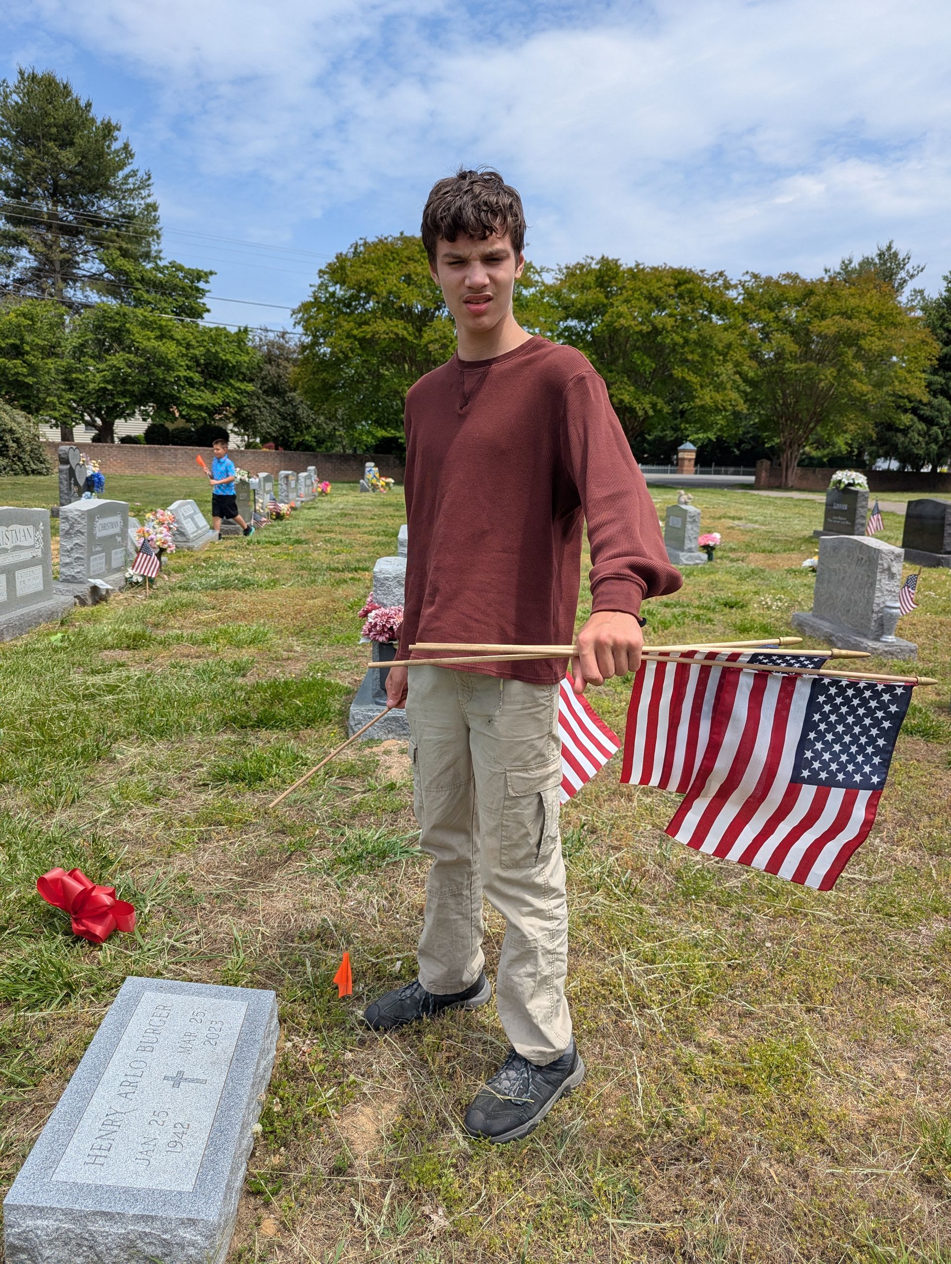 A teen standing in a cemetery beside a small American flag and gravestone, holding a string or rod
