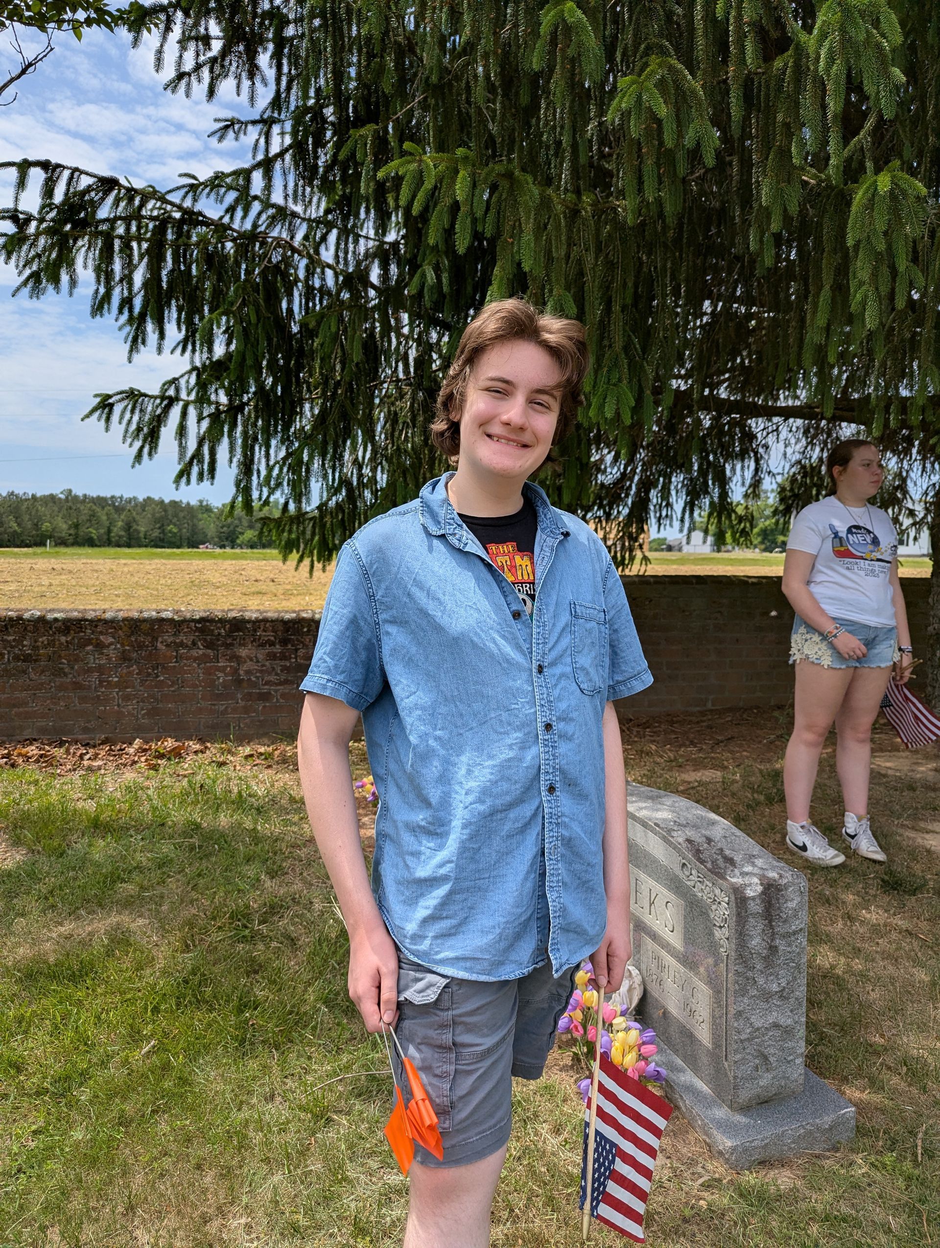 A teen smiling outdoors under a tree, holding a small orange item and an American flag bag.