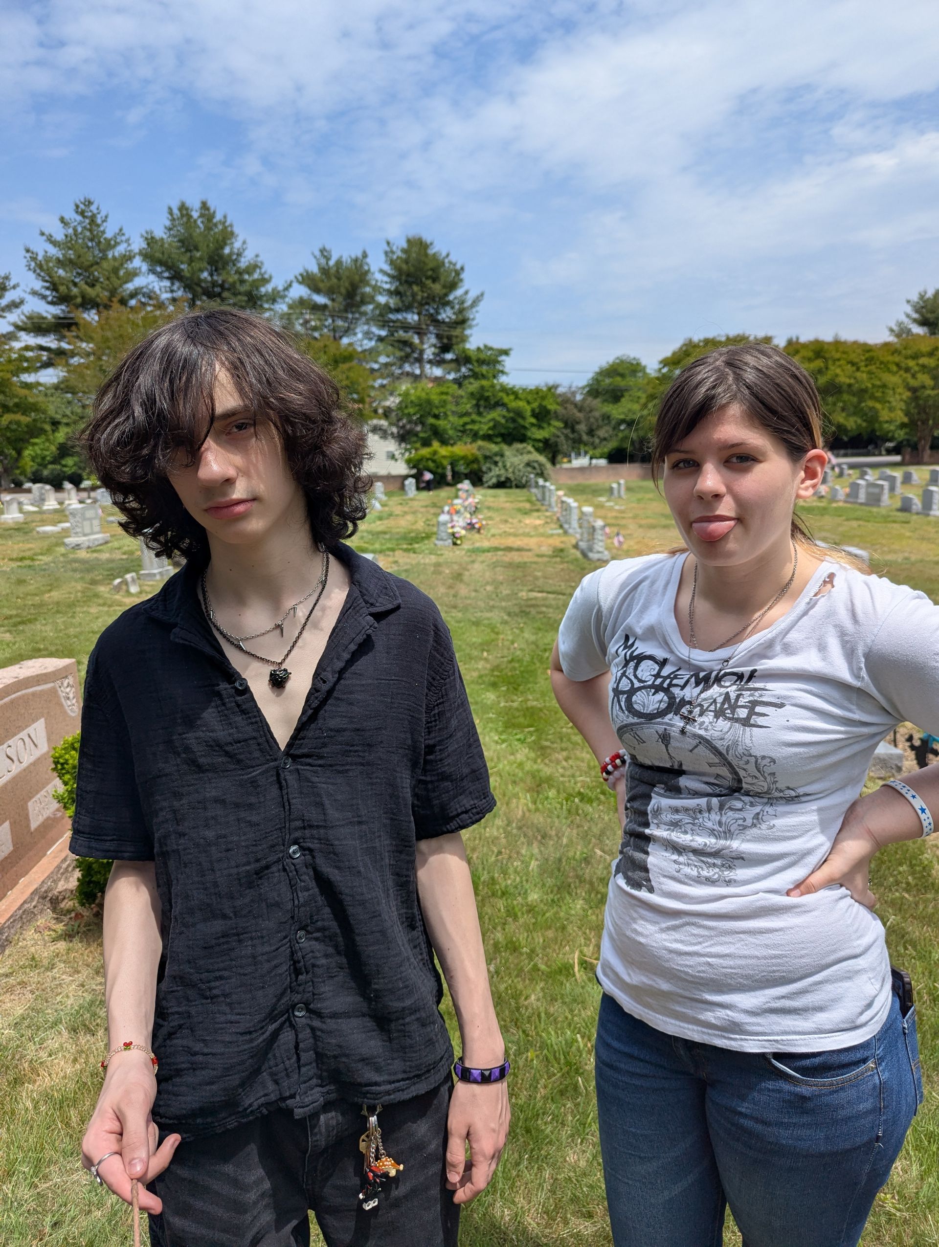 Two teens standing in a cemetery, one in black and one in a gray T-shirt, under a cloudy sky