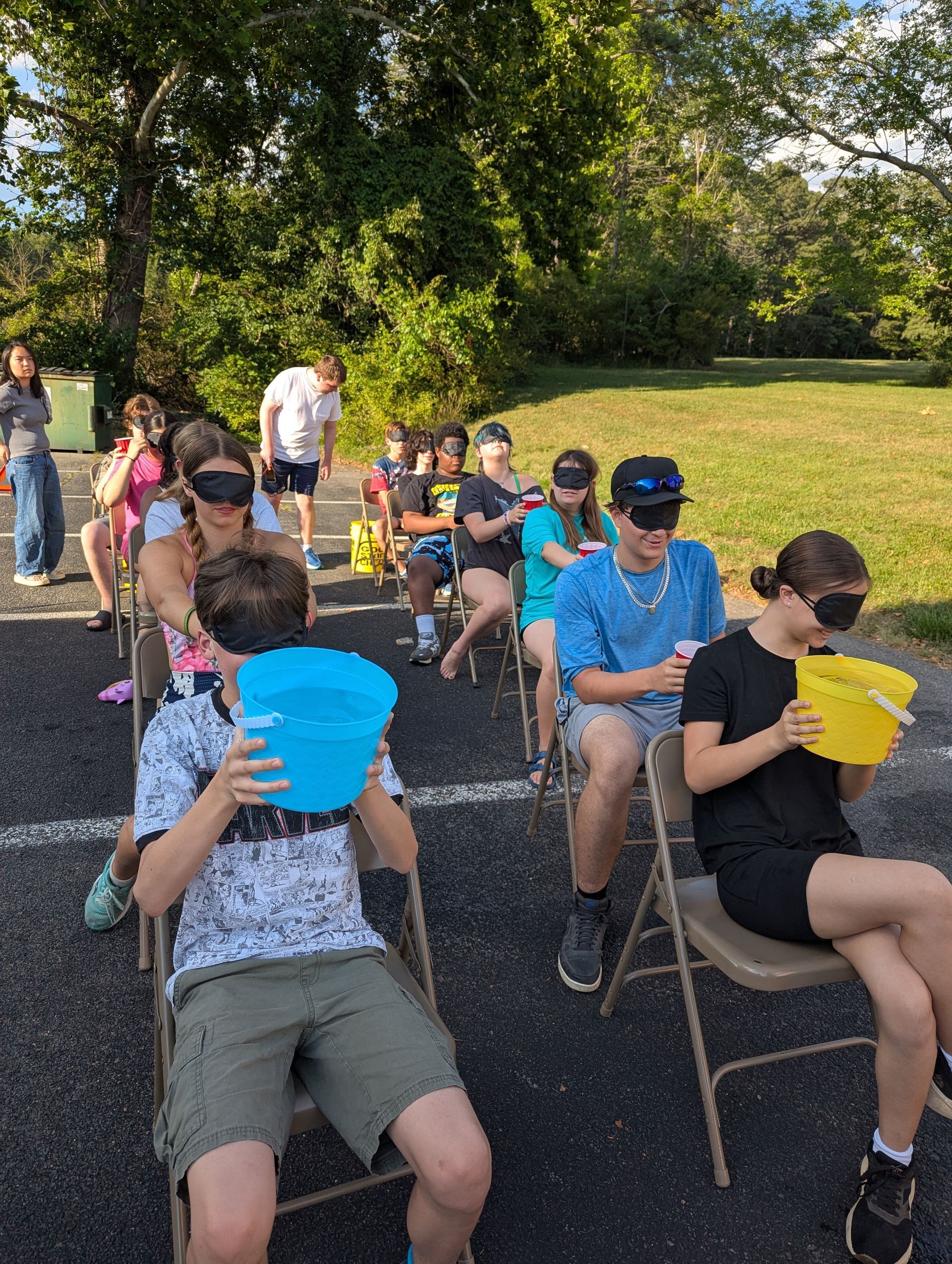 Youth playing a game outdoors in folding chairs, passing water in a cup while blindfolded
