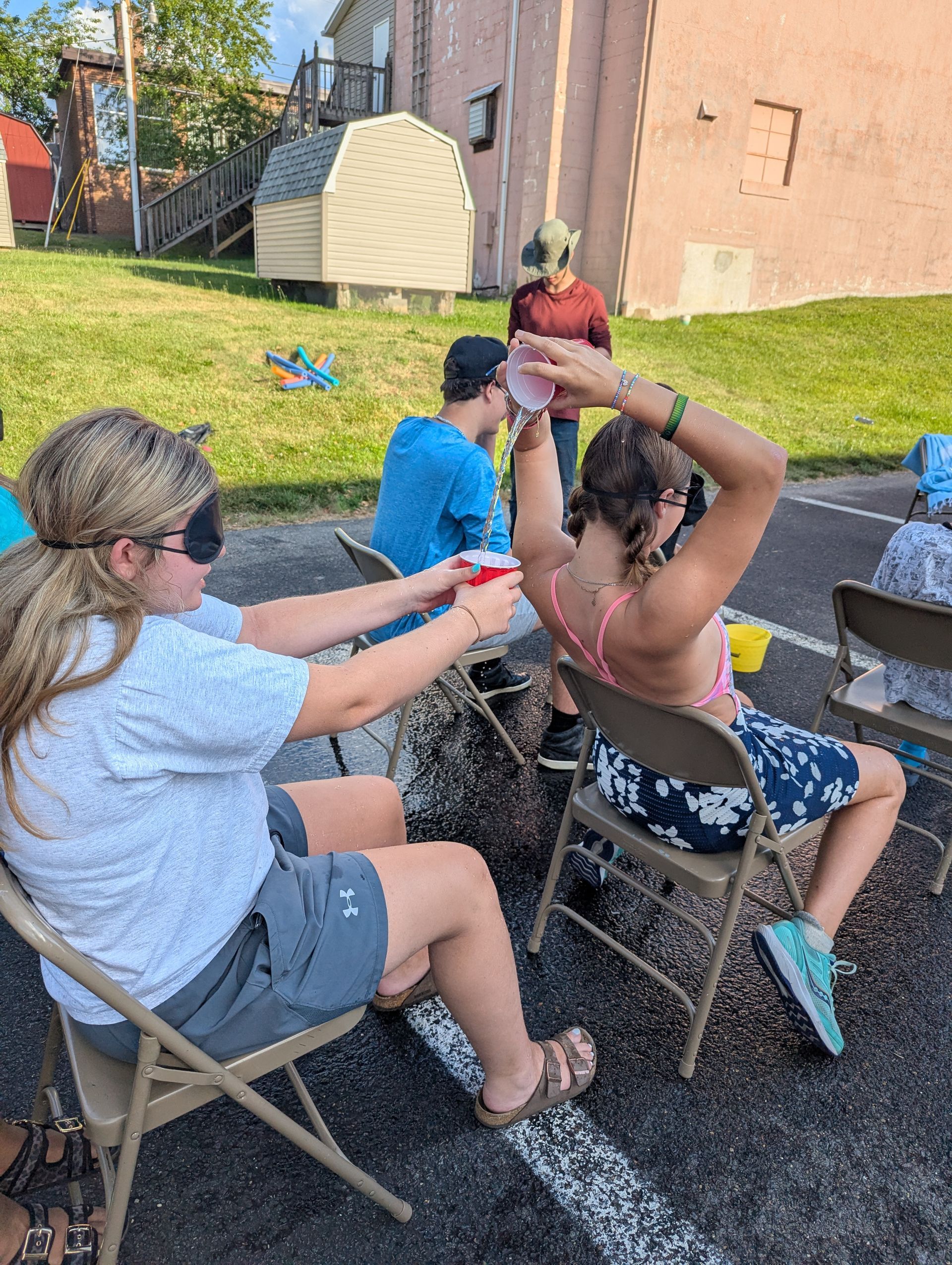Youth playing a game outdoors in folding chairs, passing water in a cup while blindfolded