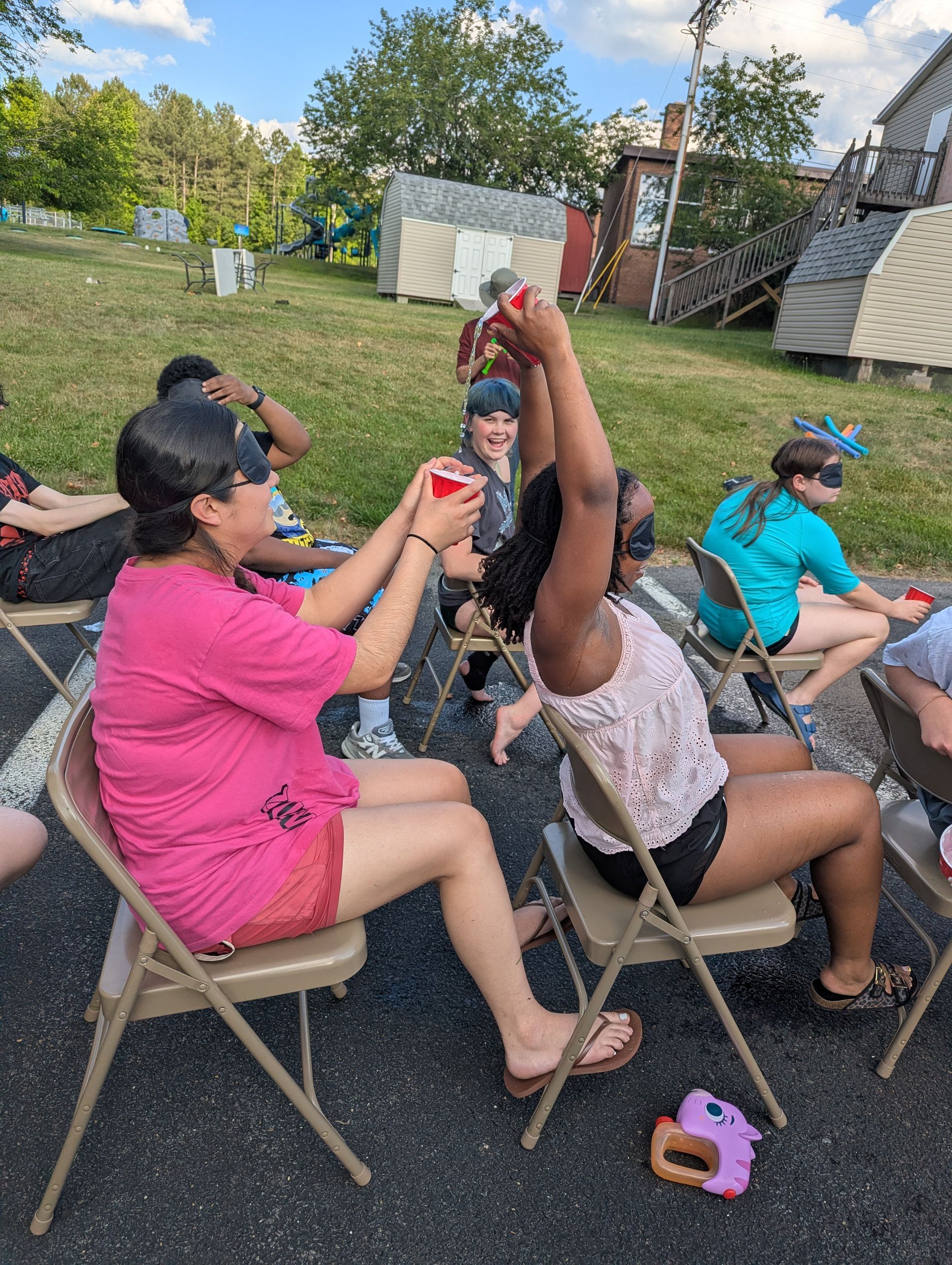 Youth playing a game outdoors in folding chairs, passing water in a cup while blindfolded