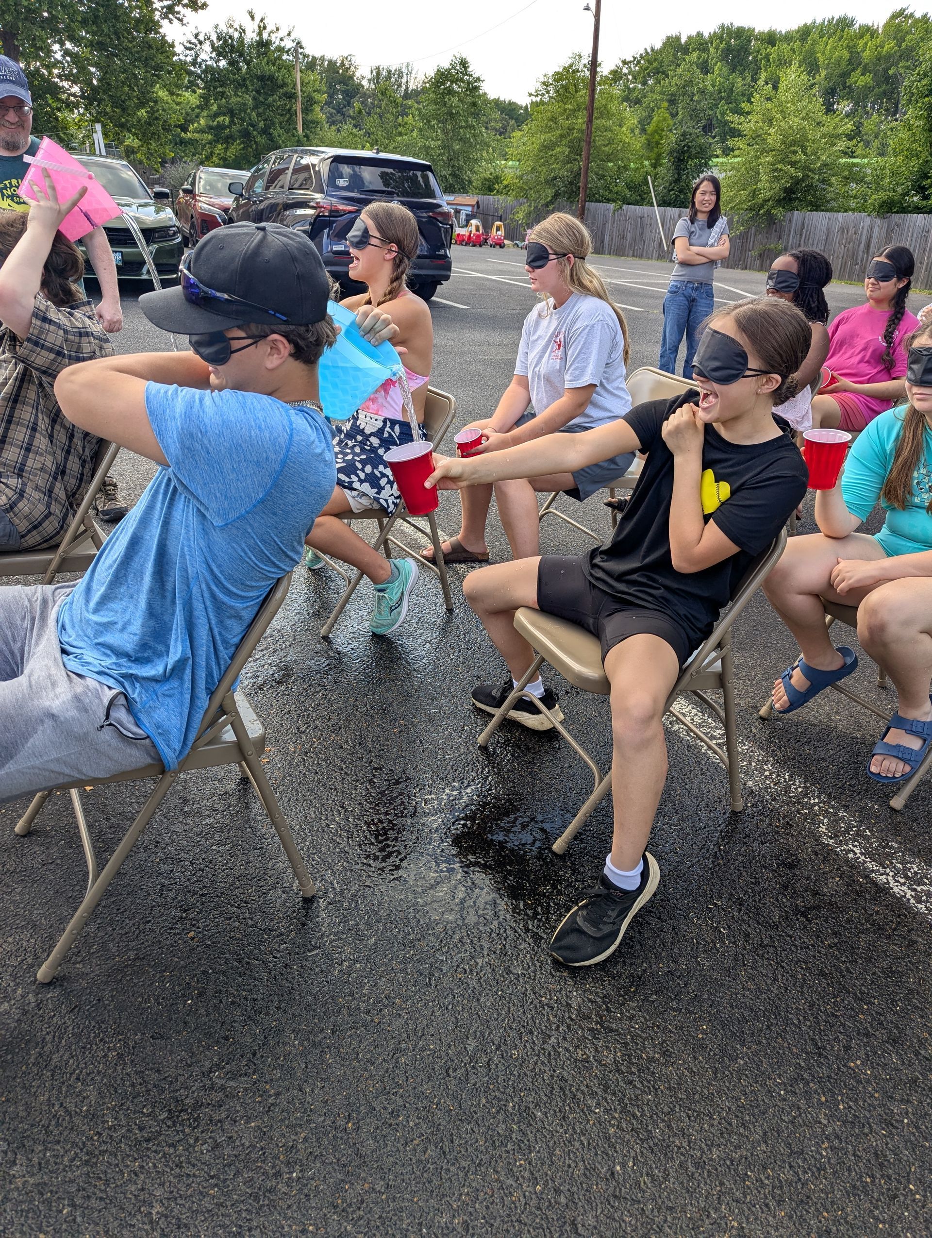 Youth playing a game outdoors in folding chairs, passing water in a cup while blindfolded