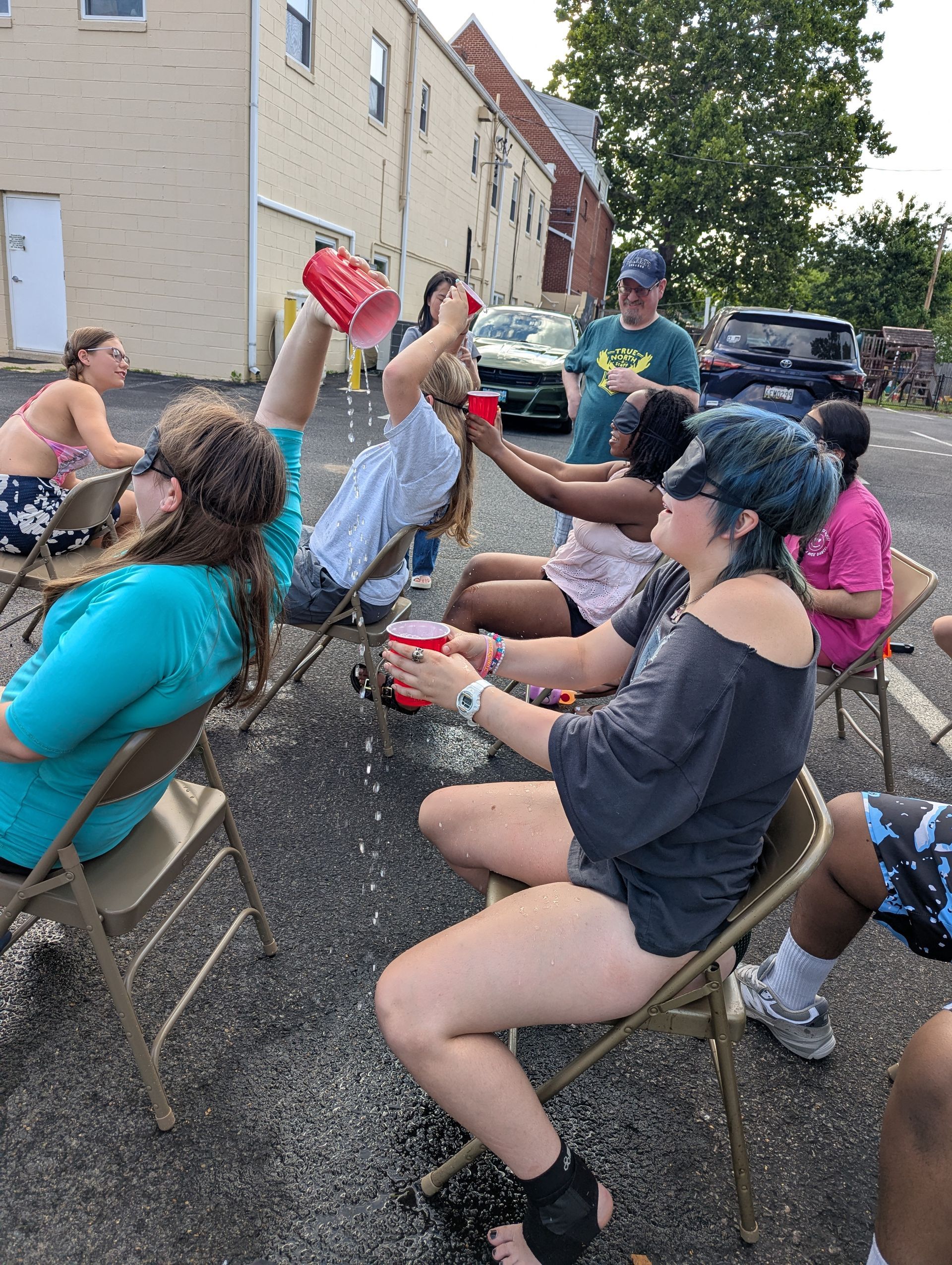Youth playing a game outdoors in folding chairs, passing water in a cup while blindfolded