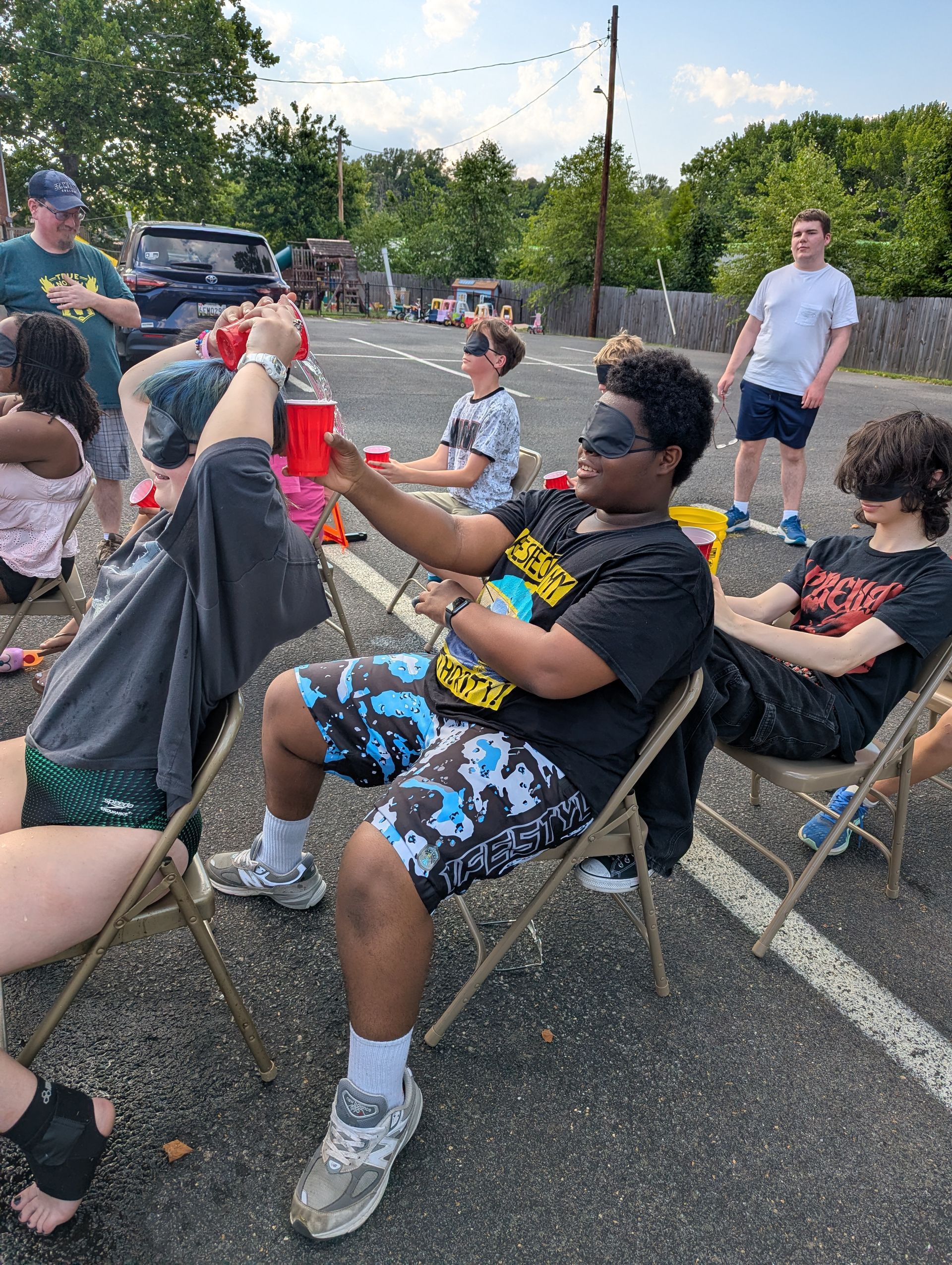 Youth playing a game outdoors in folding chairs, passing water in a cup while blindfolded