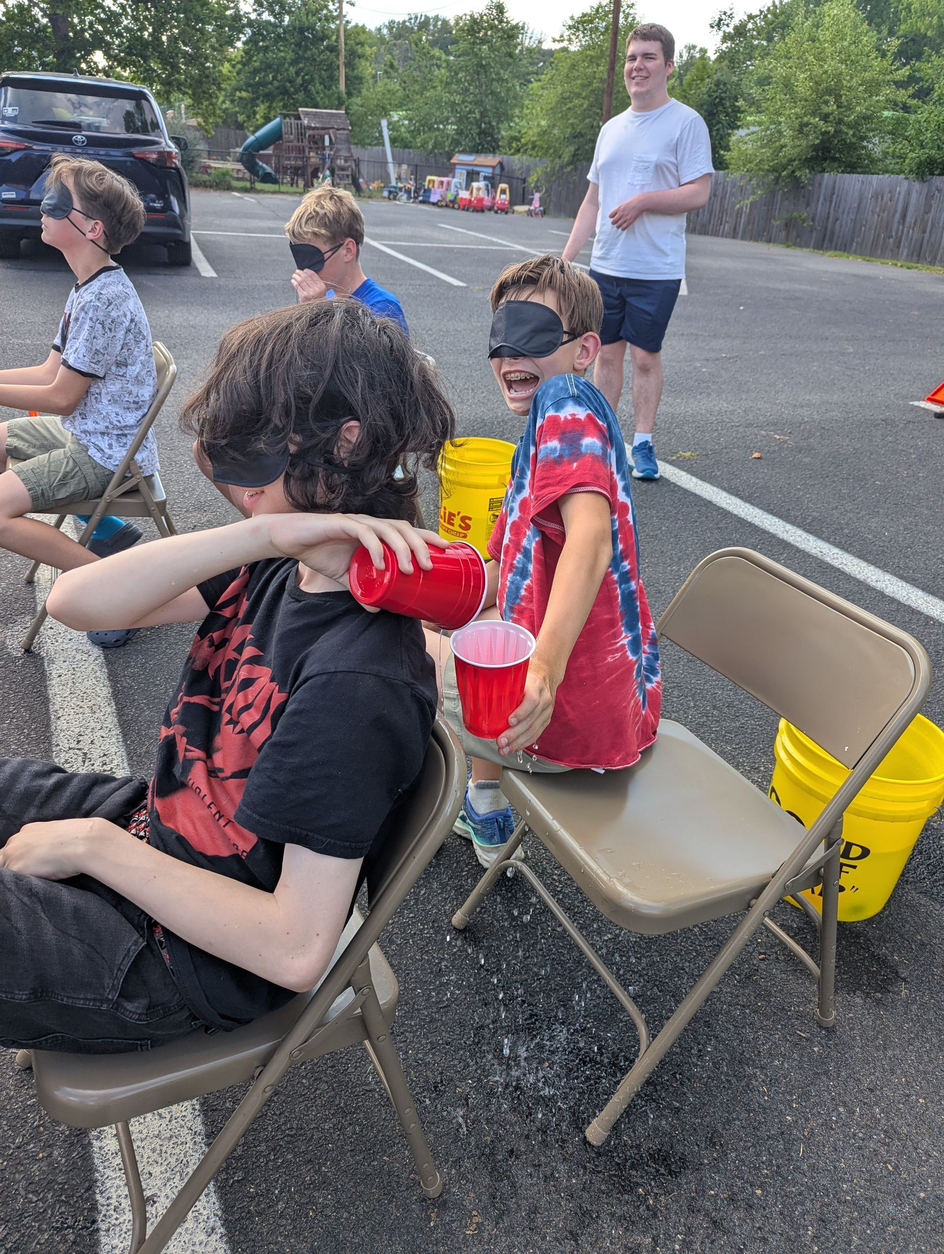 Youth playing a game outdoors in folding chairs, passing water in a cup while blindfolded, one youth moving away from the pouring water