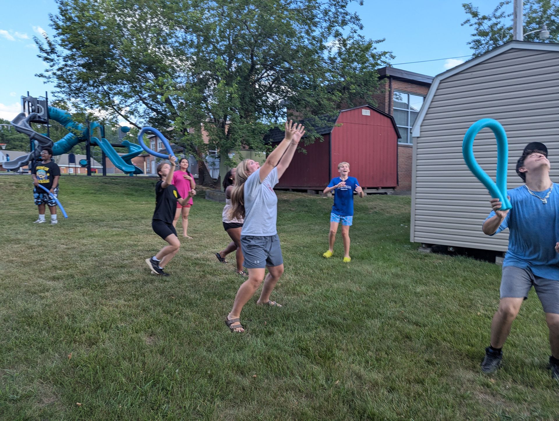 Youth playing with pool noodles on a grassy field near playground equipment and a shed.