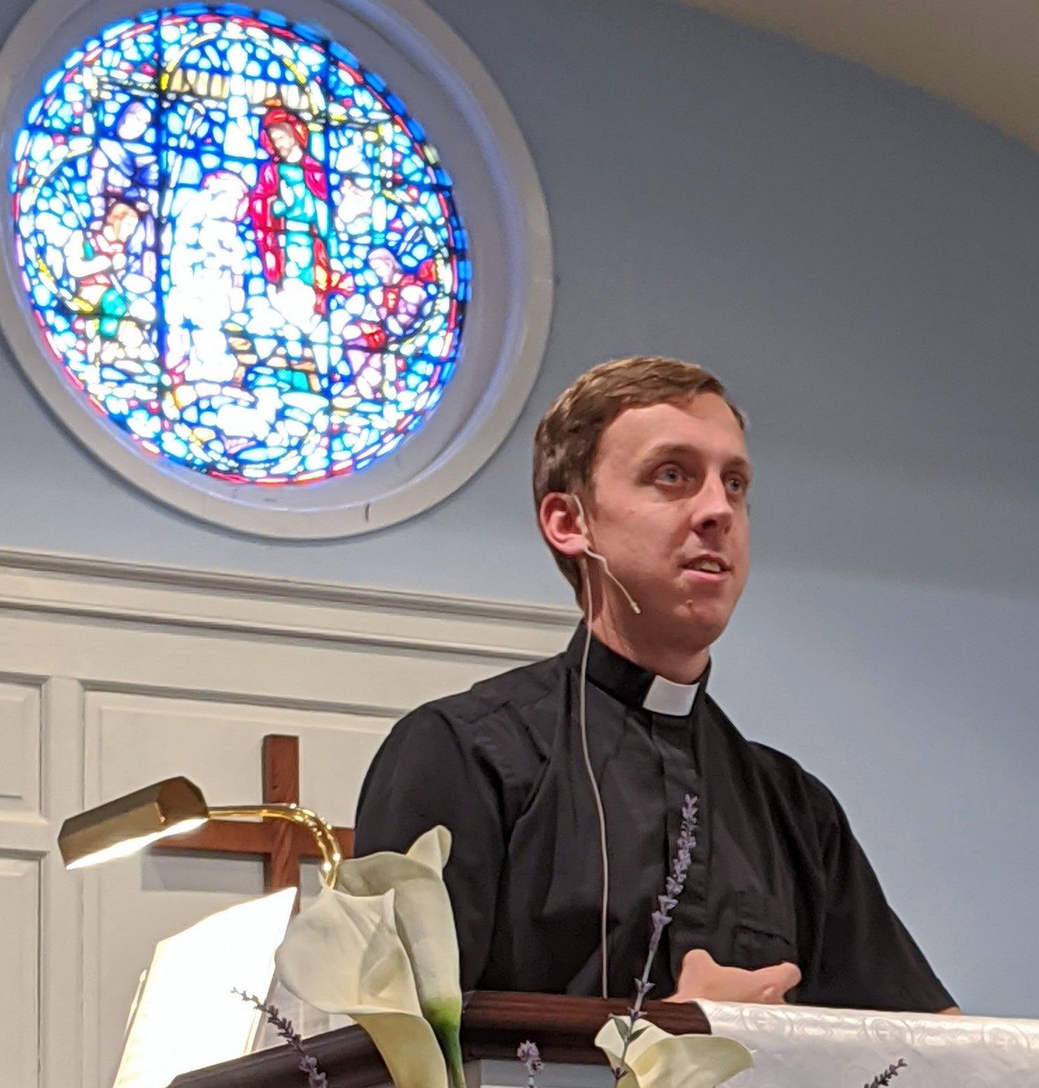 A man in a clerical collar speaks at a lectern in front of a stained-glass window.