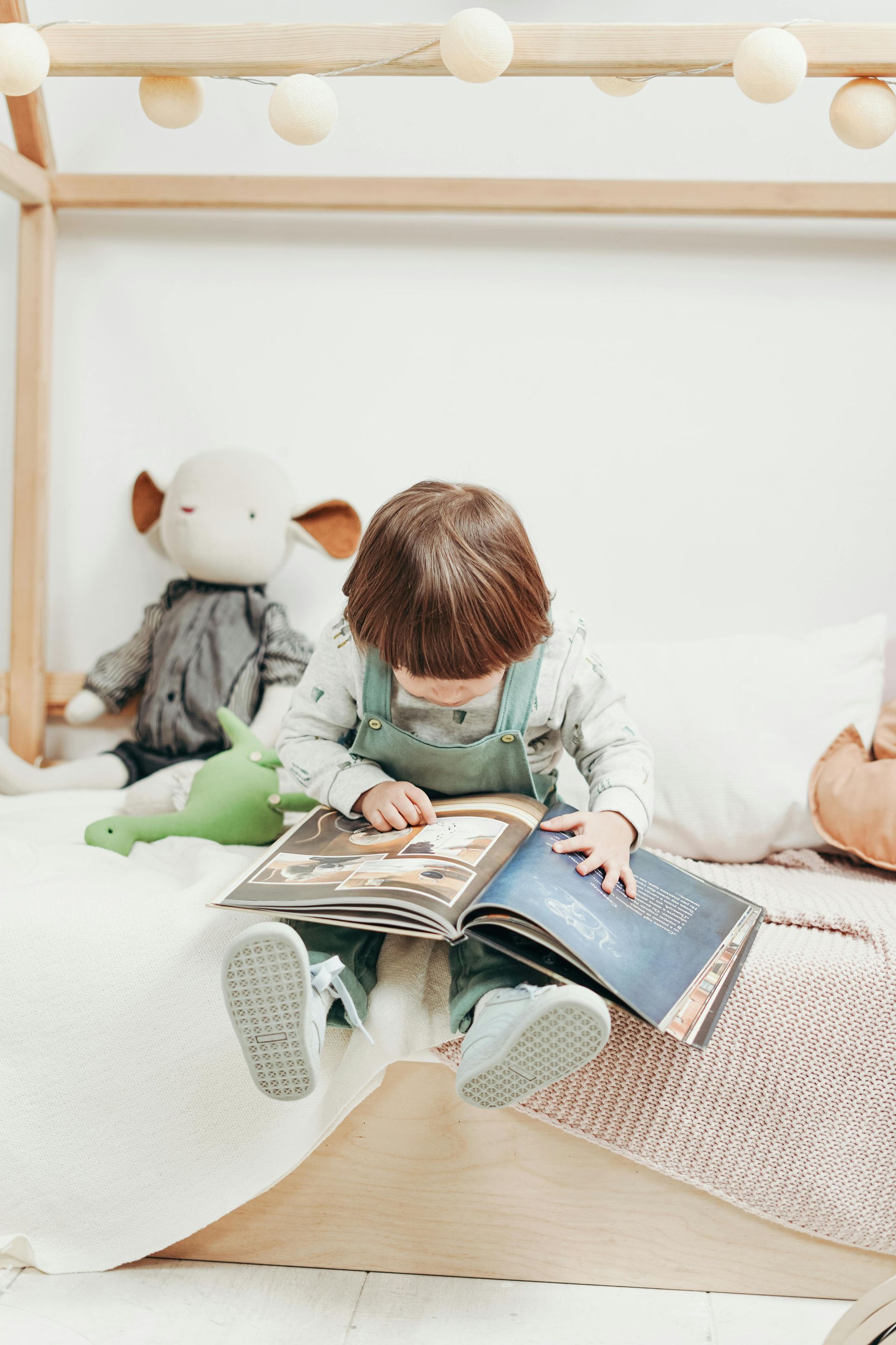 Child in overalls reading a book on a bed with stuffed toys; string lights overhead.