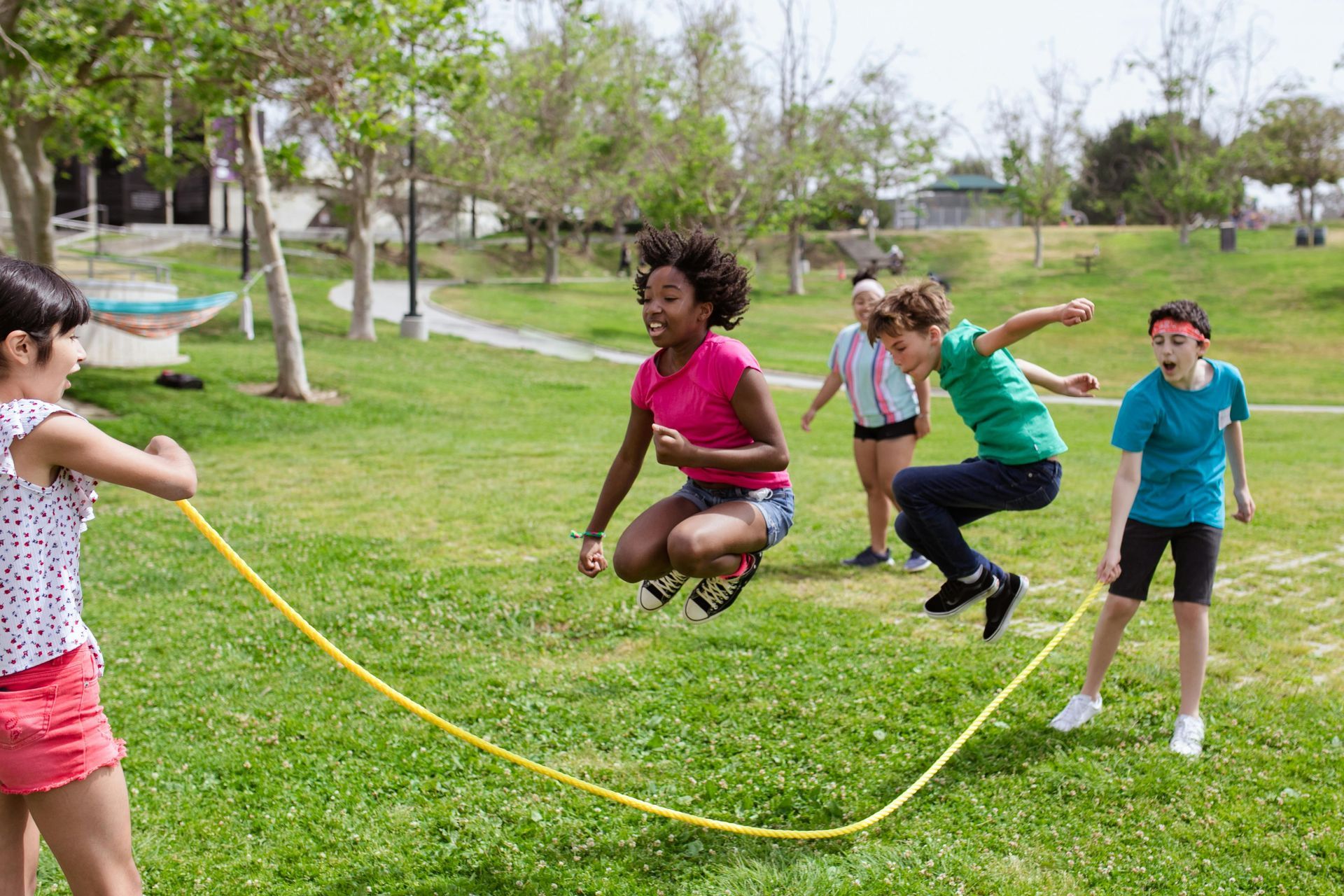 Children playing jump rope in a grassy park on a sunny day.