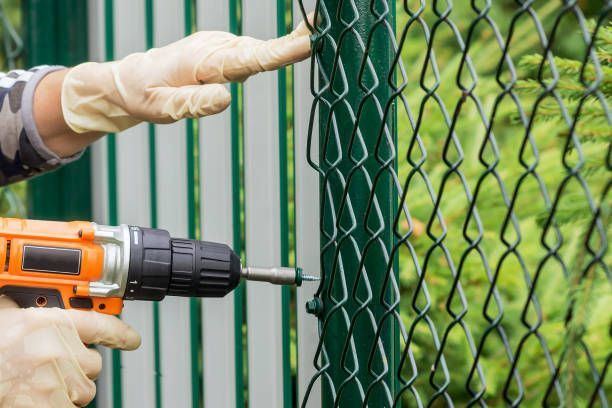 A worker using an orange drill to fasten a green metal fence outdoors.