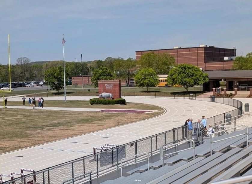 Workers installing a dark metal fence panel with a power drill a school athletic complex with a running track, football field, and spectator bleachers.