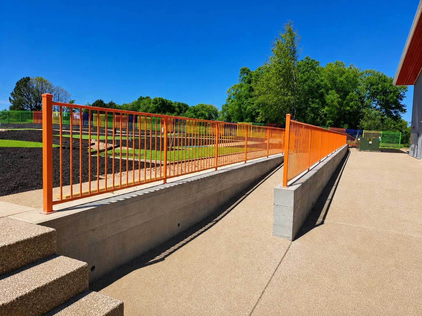 Workers installing a dark metal fence panel with a power drill an accessible concrete ramp and walkway with bright orange safety railings.