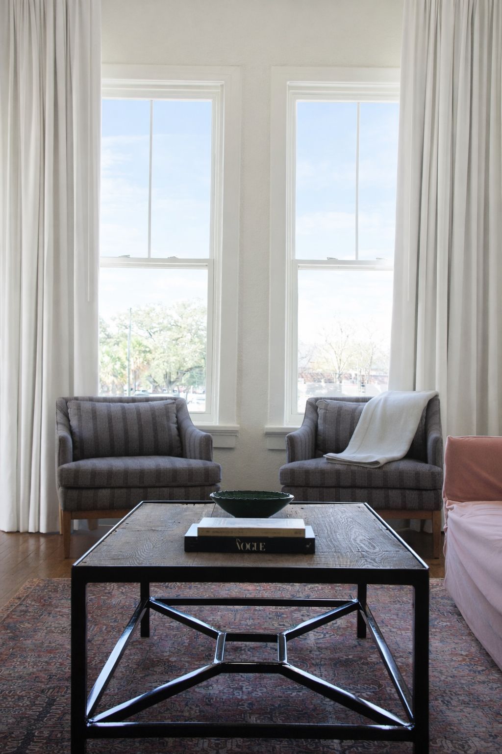 A coffee table sits between two gray armchairs in front of tall windows with long white curtains.