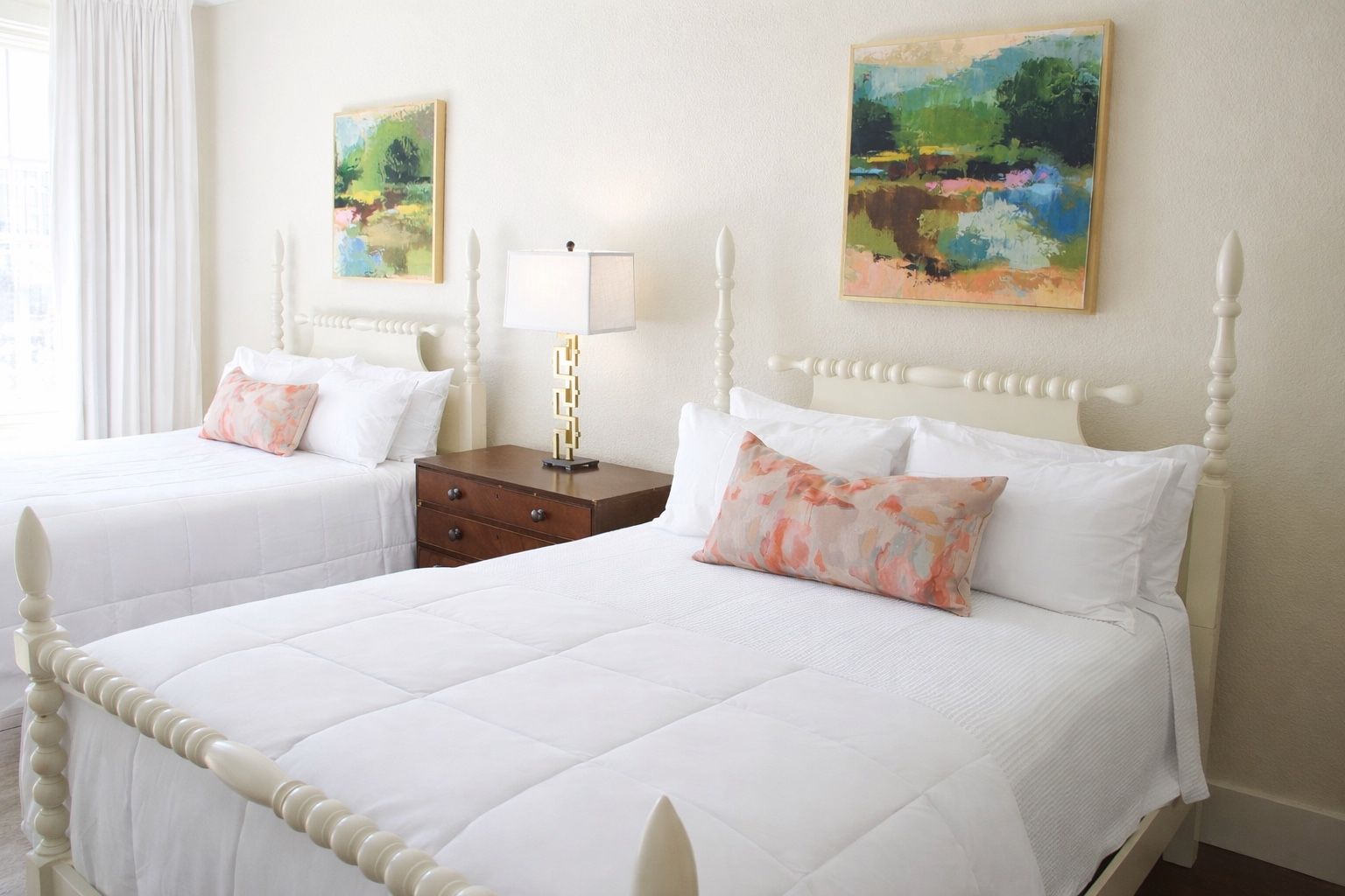 Two beds with white bedding, floral accent pillows, and landscape paintings in a brightly lit, neutral-toned bedroom.
