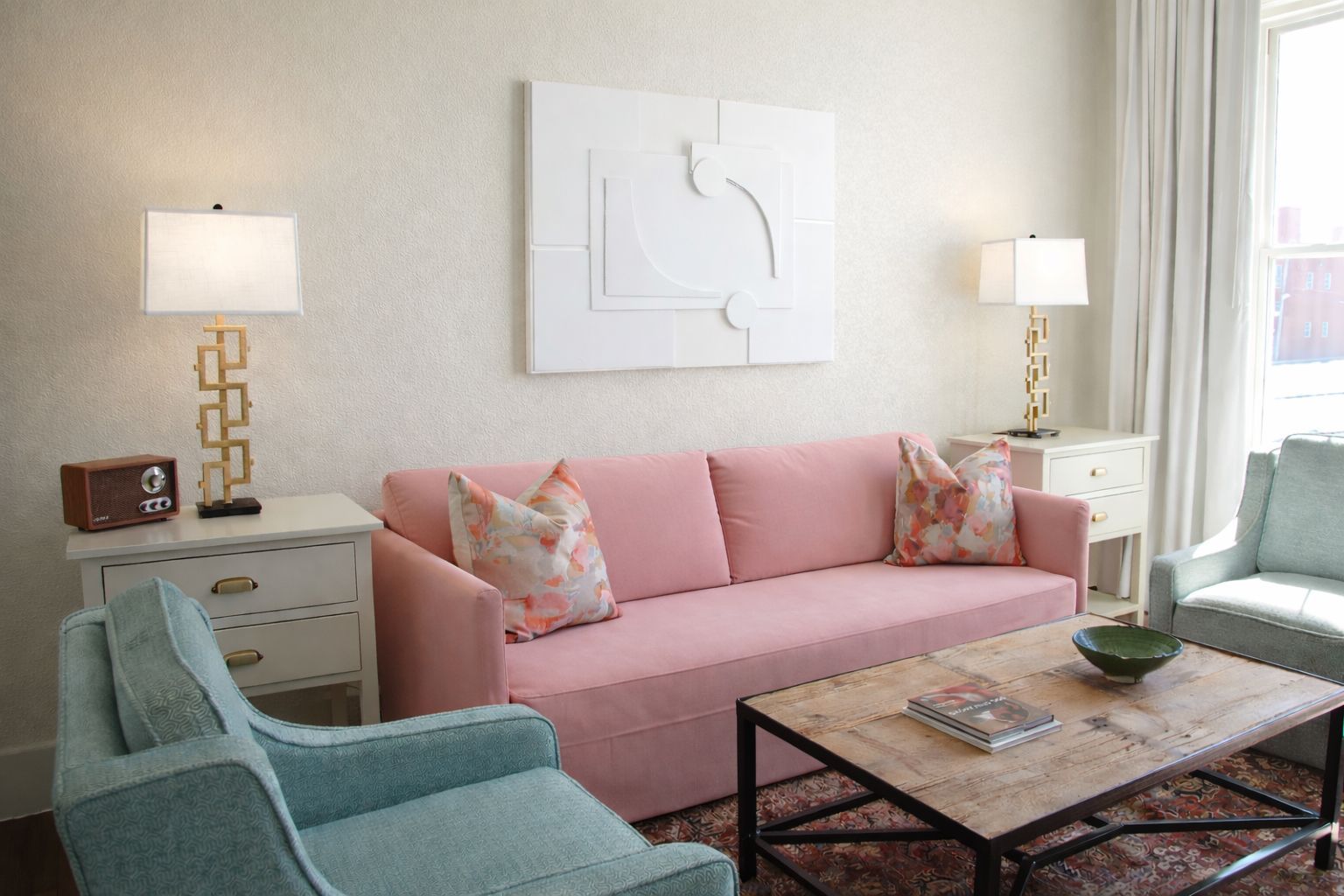A living room with a pink sofa, two teal armchairs, a wooden coffee table, and two gold lamps on white side tables.
