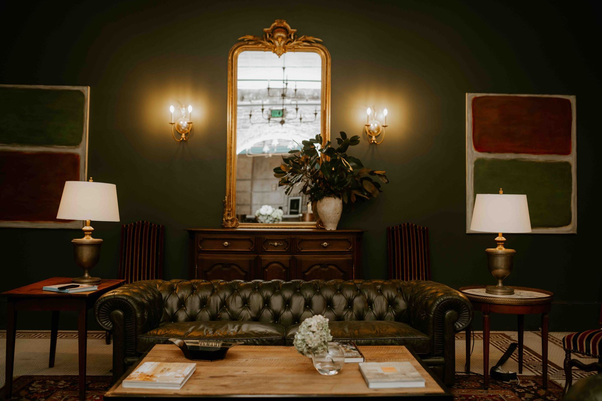 A dark green room with a tufted sofa, wooden coffee table, matching table lamps, and a central gold-framed mirror.
