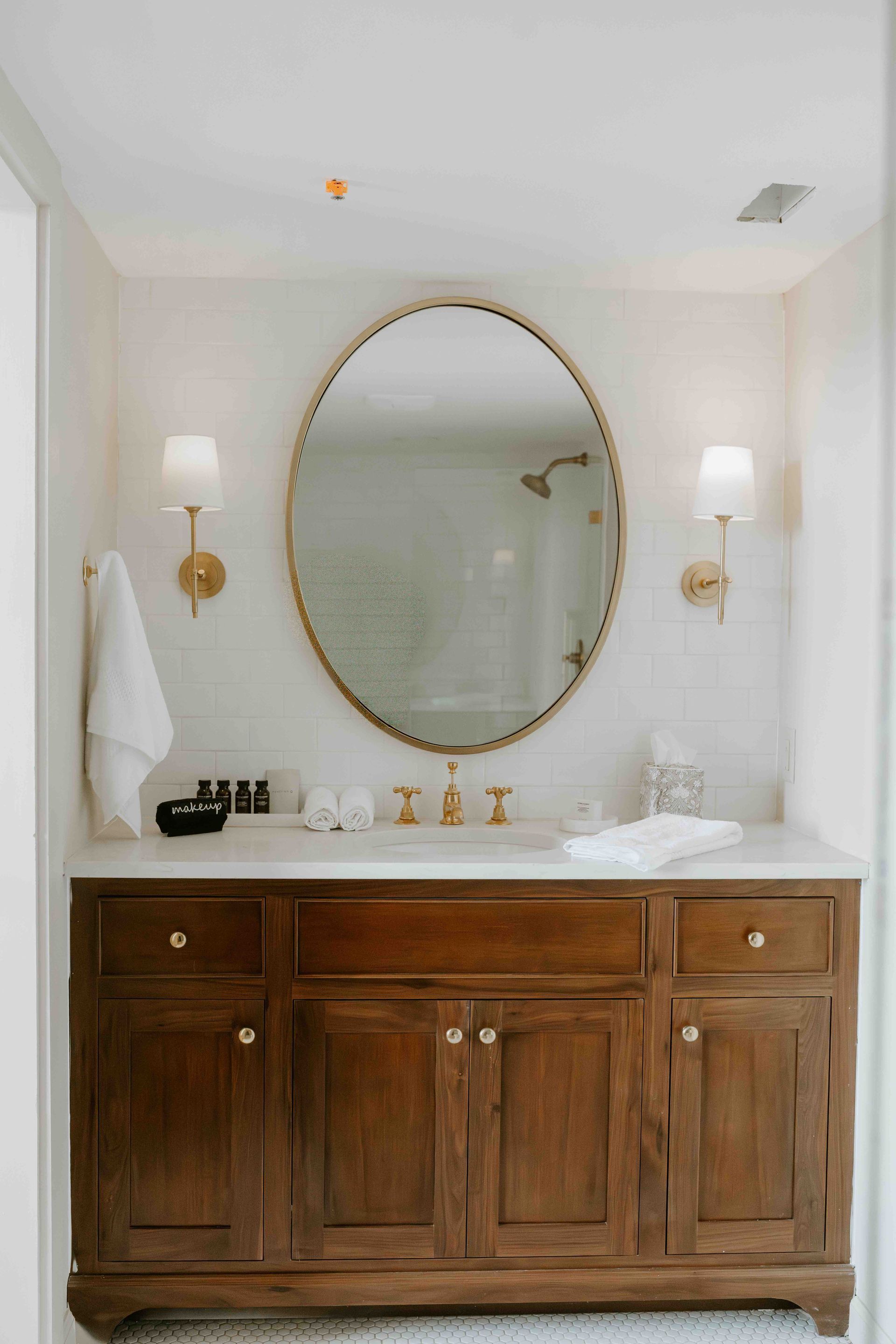 A bathroom vanity with a dark wood cabinet, white countertop, oval mirror, and gold fixtures against white subway tiles.