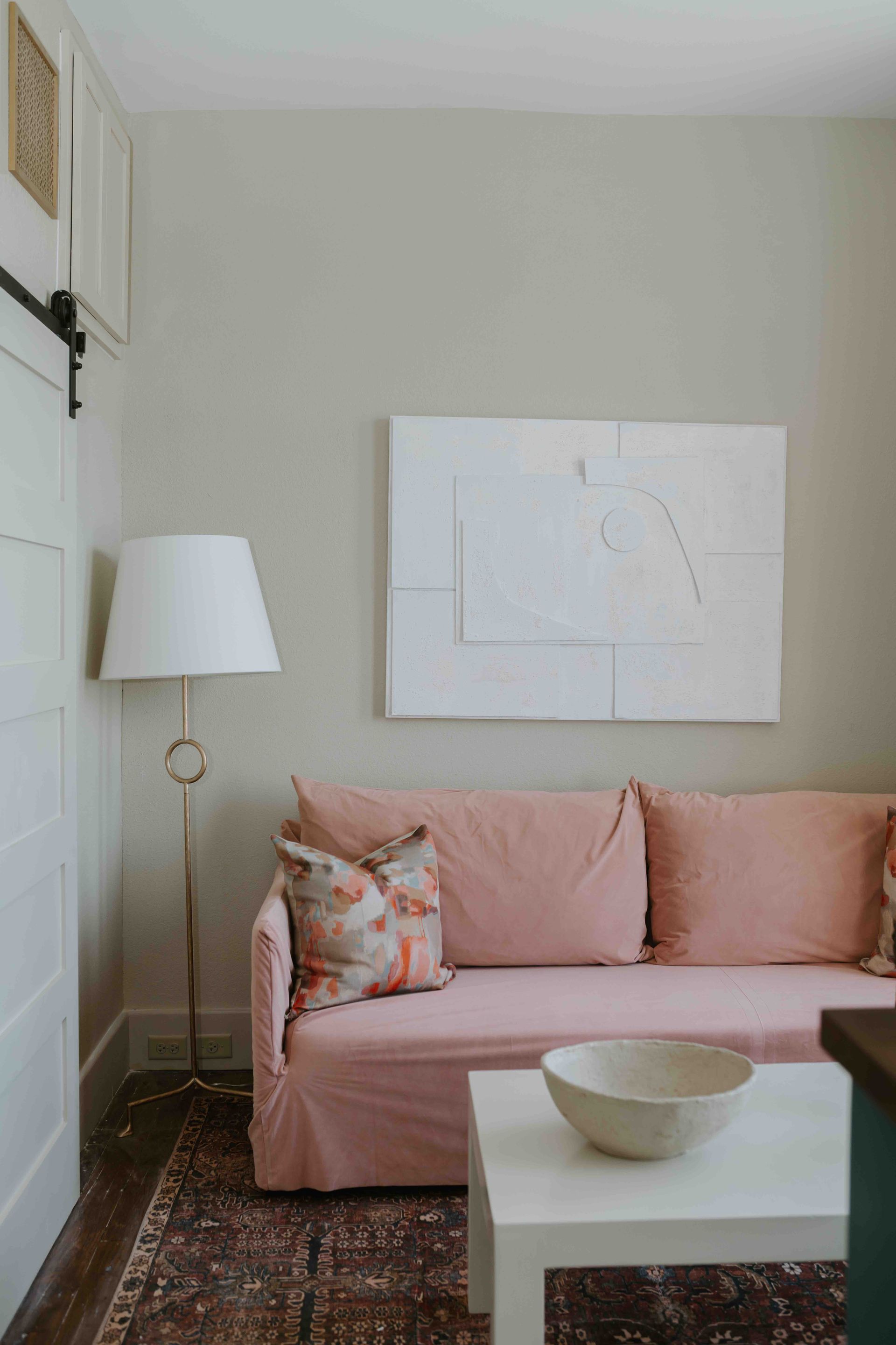 A pink couch with a patterned pillow sits next to a white lamp and a coffee table with a bowl on a vintage rug.