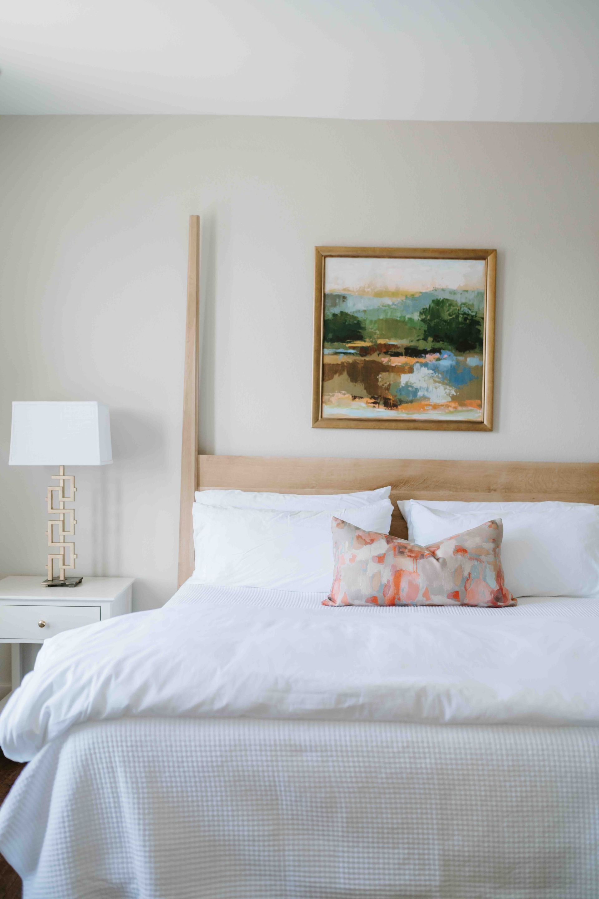 A bedroom featuring a bed with a light wood headboard, white bedding, a decorative pillow, and a landscape wall painting.