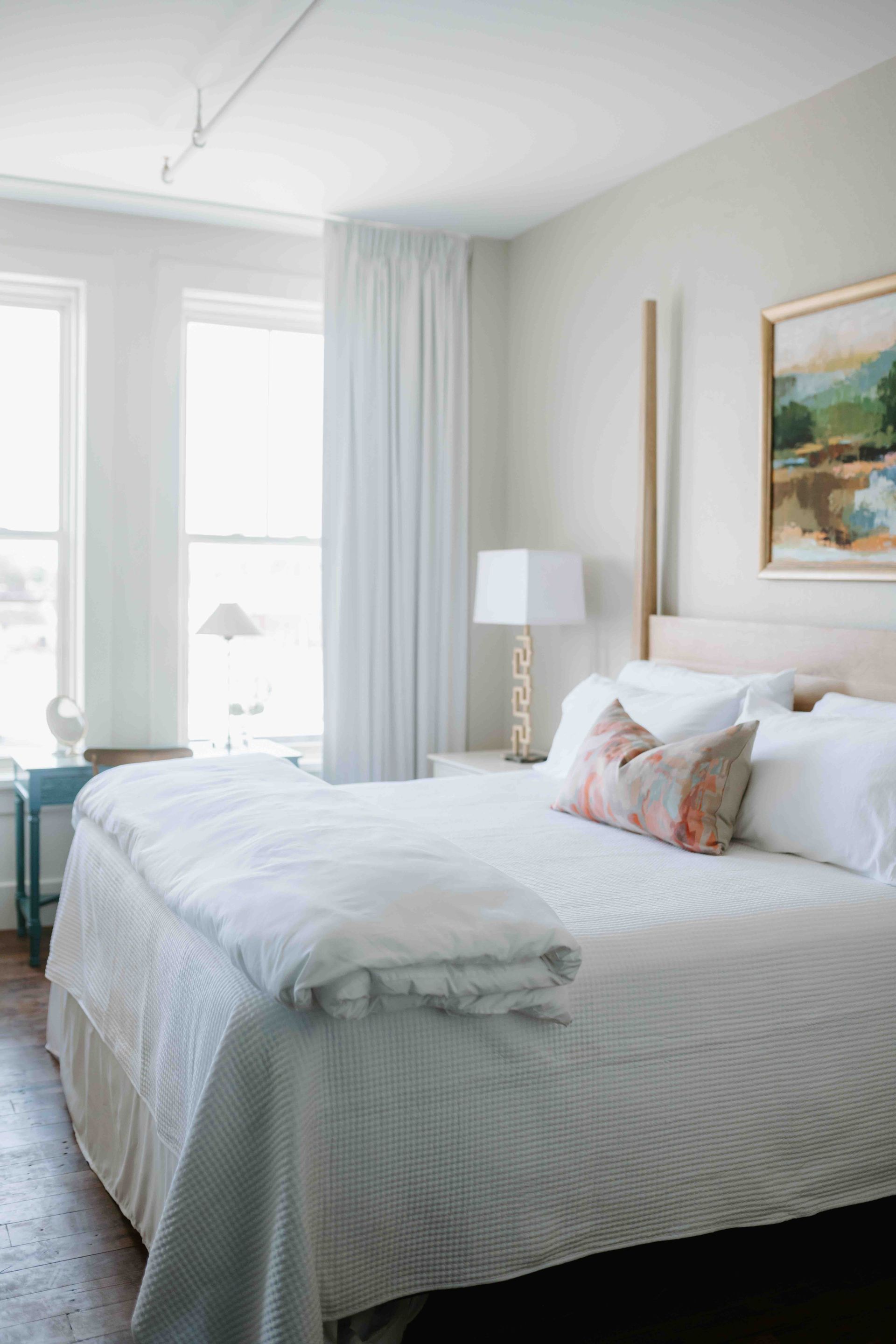 A bright bedroom featuring a queen bed with white bedding, a decorative floral pillow, and landscape art on a beige wall.