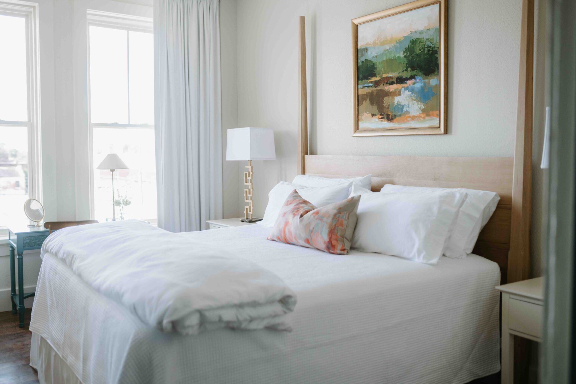 A bright bedroom featuring a queen bed with white bedding, a decorative accent pillow, and a landscape painting above.