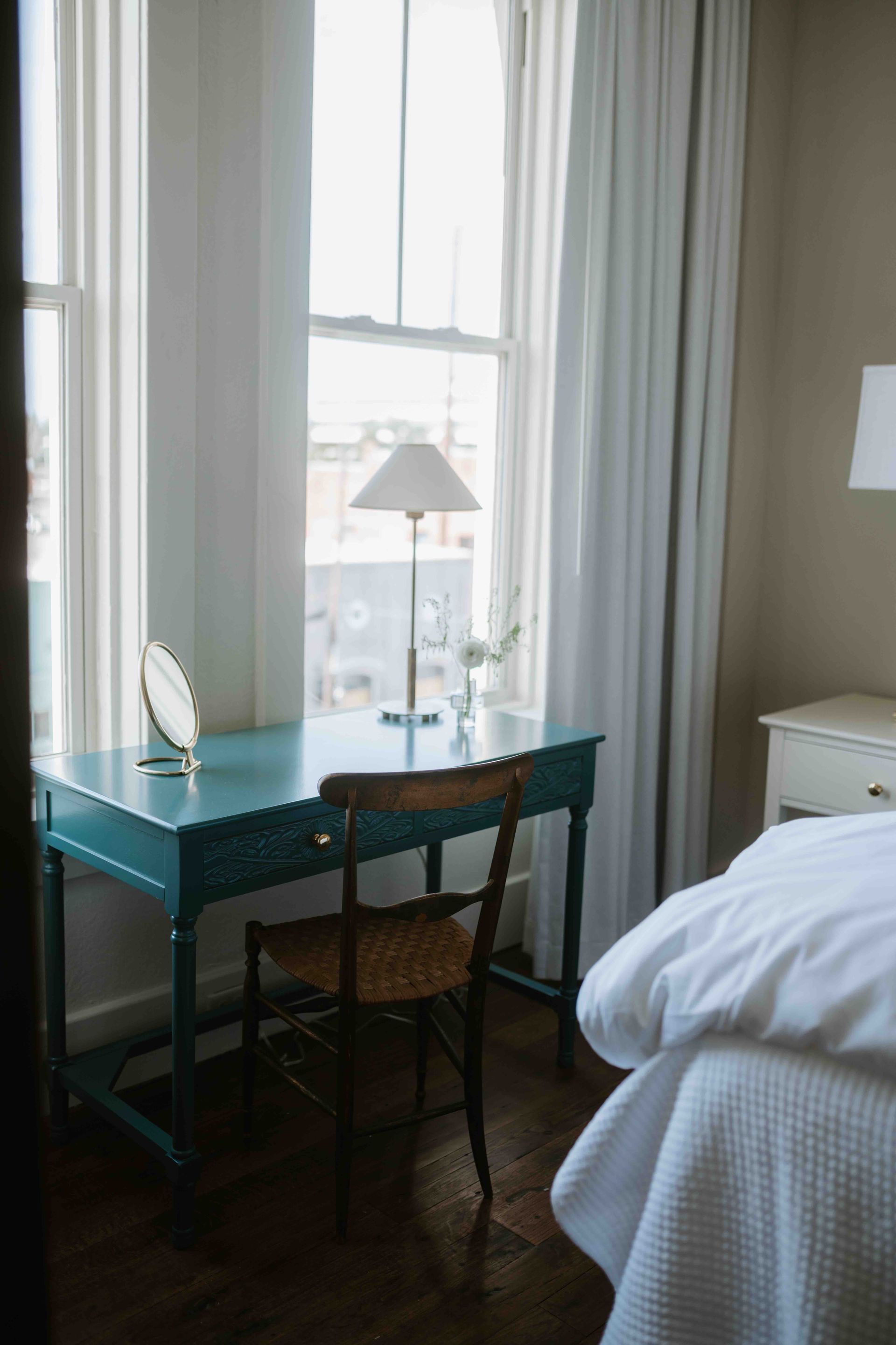 A teal wooden desk with a lamp, a small mirror, and a wooden chair in front of a bright window with white curtains.
