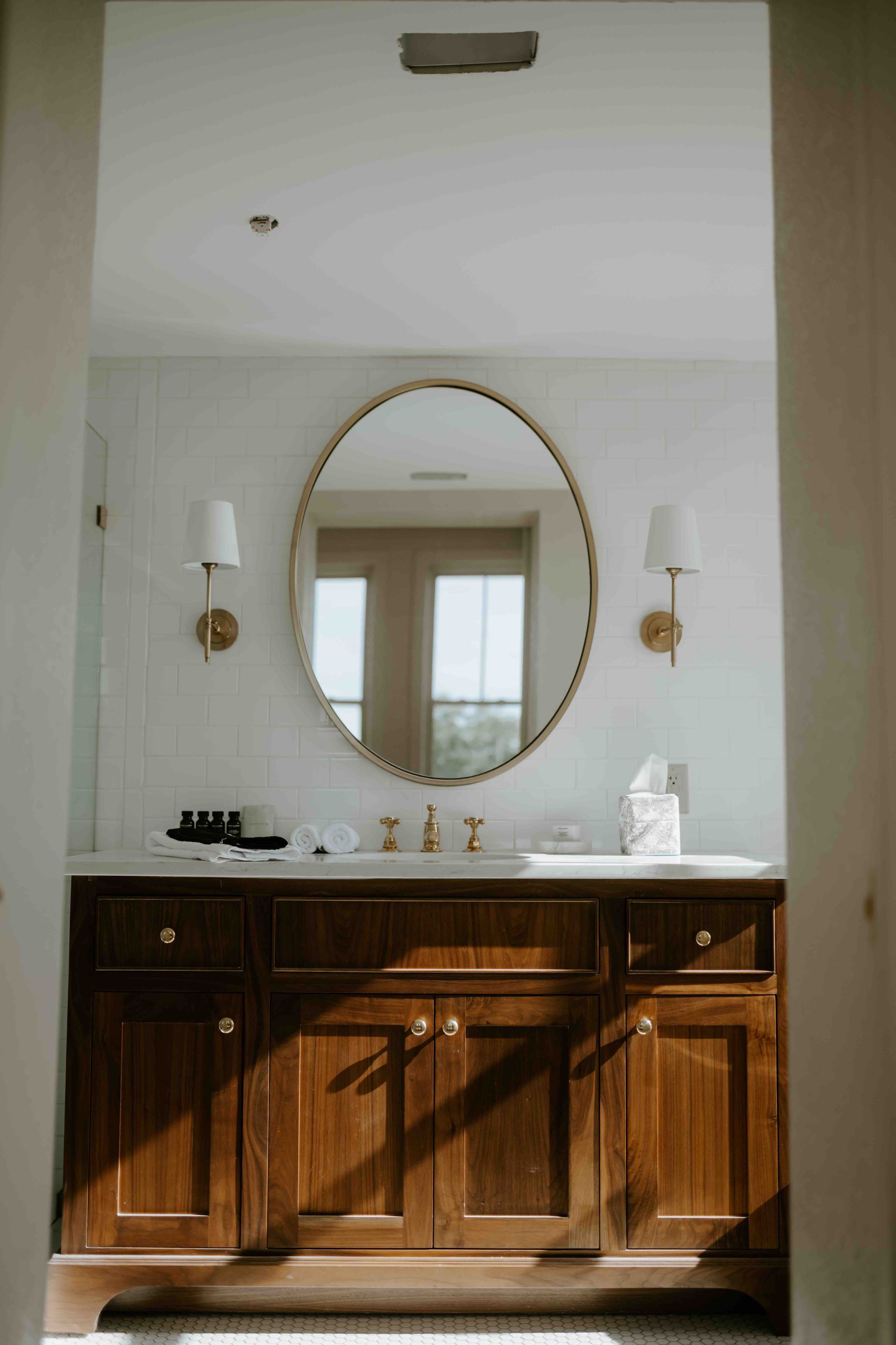 A wooden bathroom vanity with a white countertop, oval mirror, and two wall sconces set against a white wall.