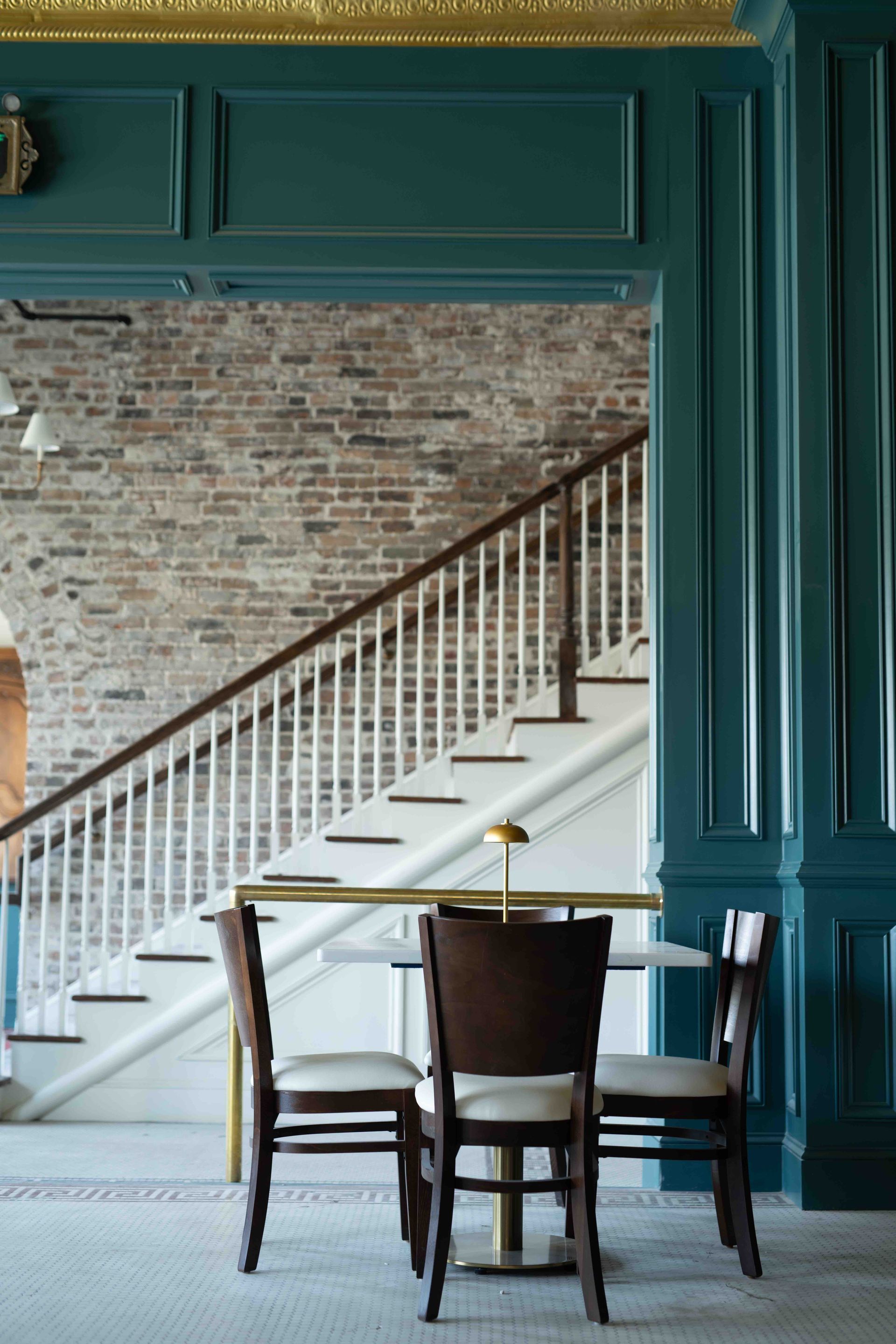 A small table with three wooden chairs set against teal paneled walls, with a staircase and brick wall in the background.