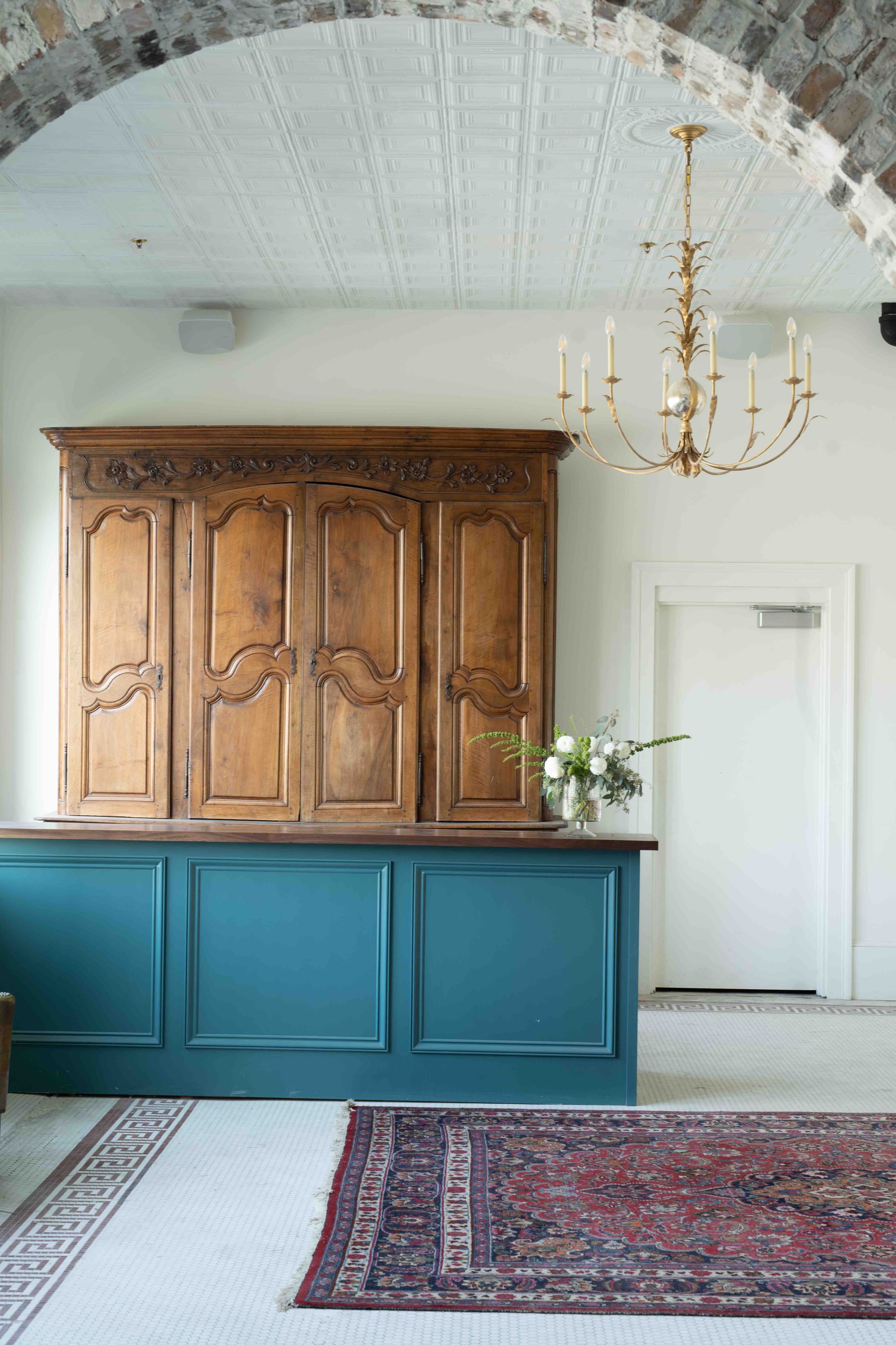 A rustic bar with a teal base and large wooden cabinet, set under a stone archway with a chandelier and red patterned rug.